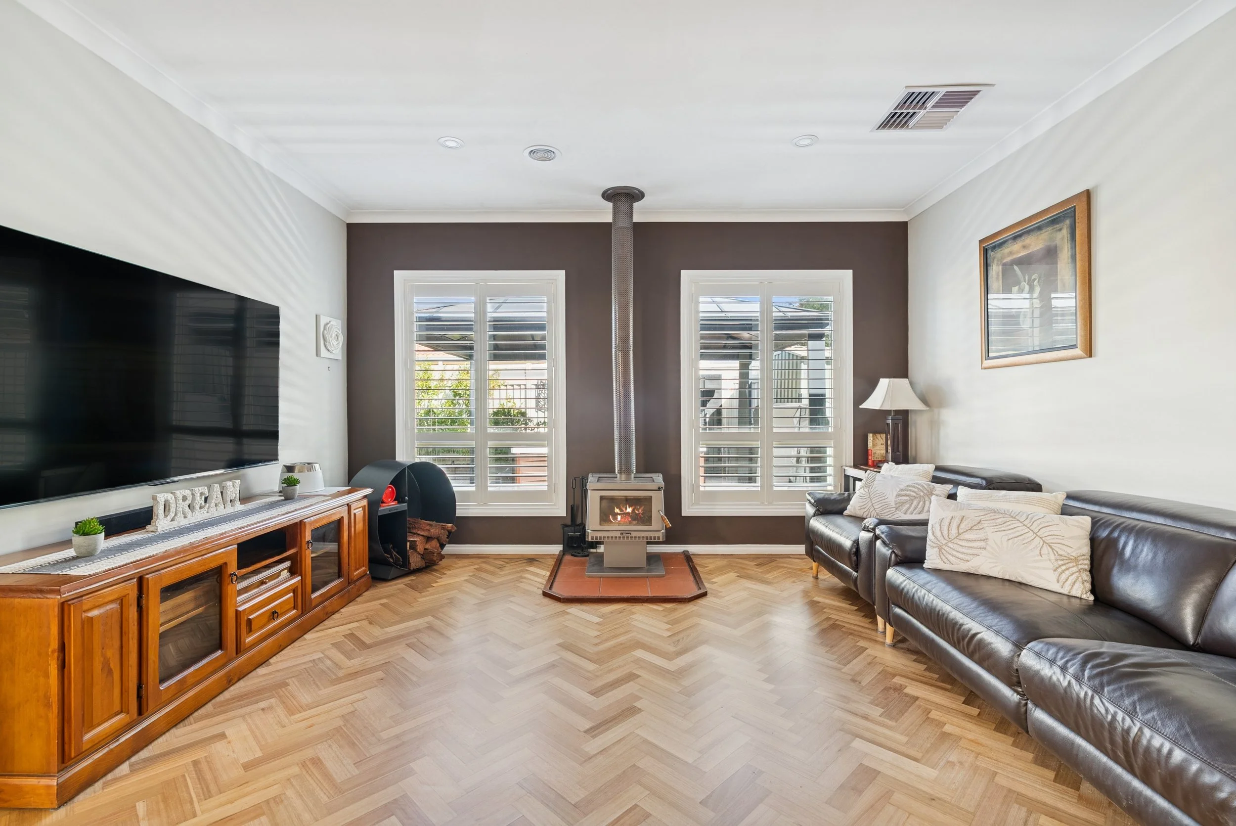 Living room with large television, brown accent wall, wood floor, black leather sofa, fireplace with chimney, windows with white blinds, side table with lamp, and framed artwork on wall.