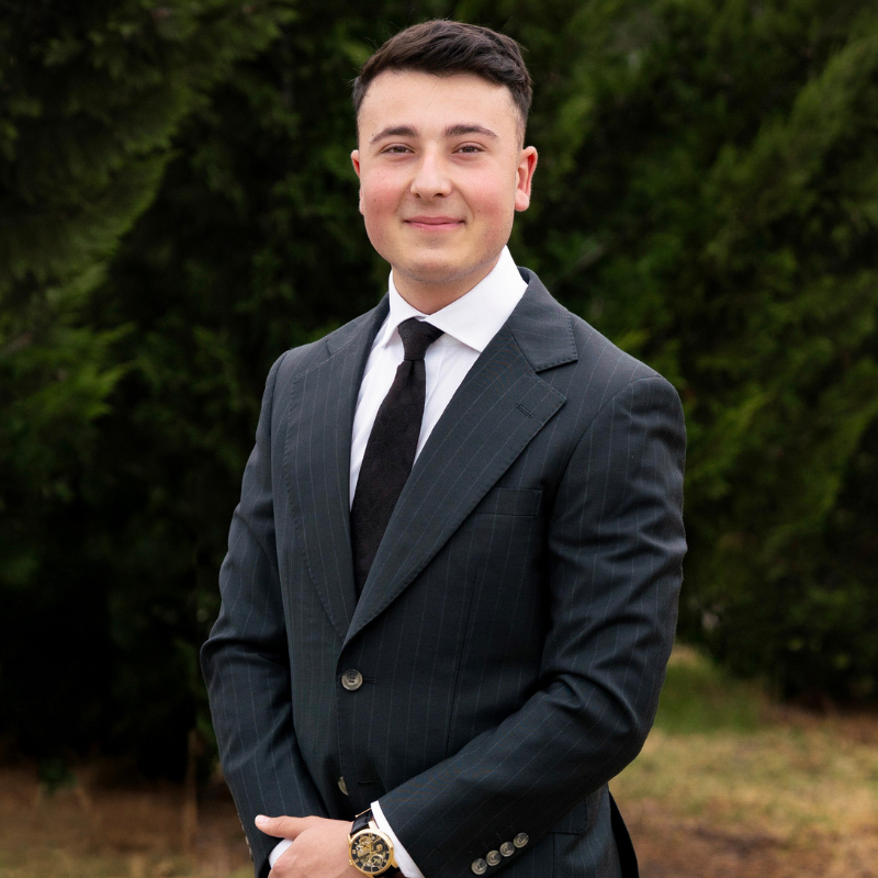 A man in a dark pinstripe suit and white shirt, smiling, against a black background.