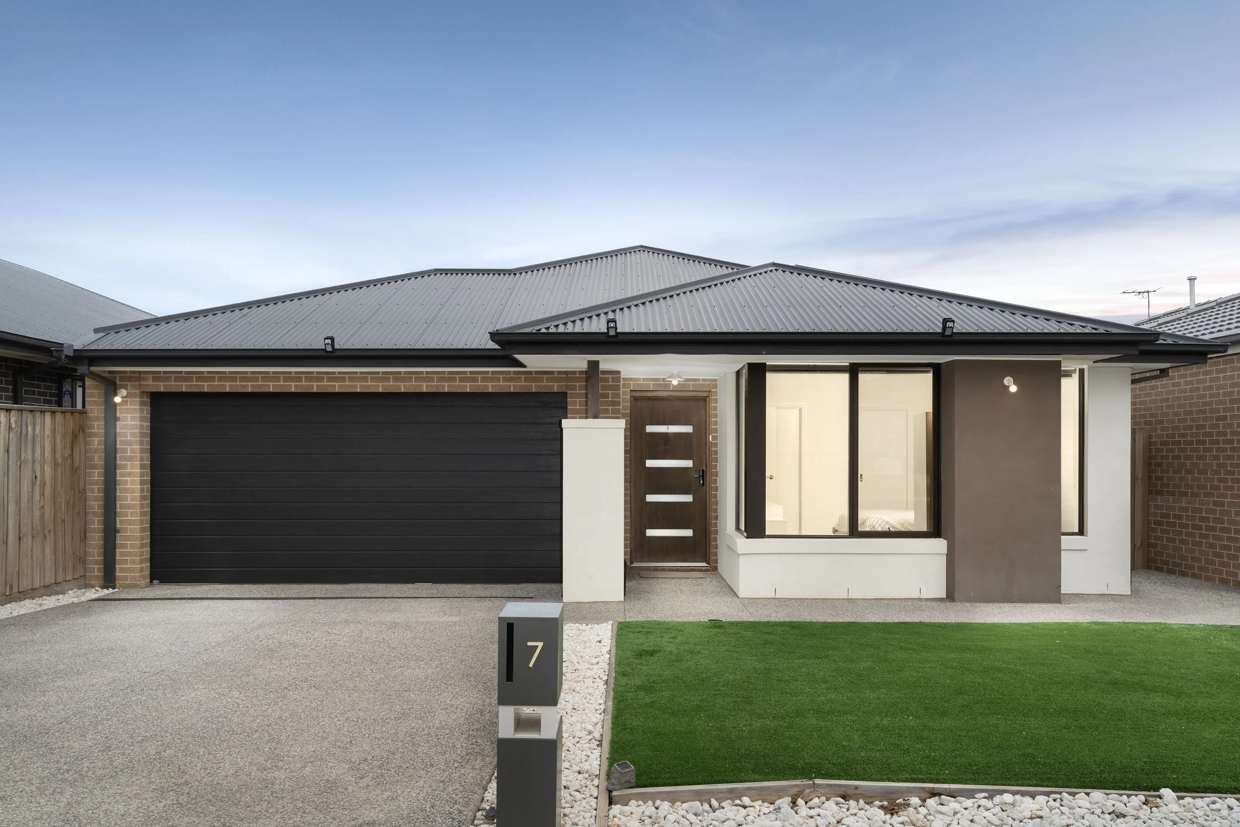 Front view of a modern house with a black garage door, brown front door with horizontal glass panels, large windows, a small front lawn, and a mailbox with the number 7, during twilight or early evening.