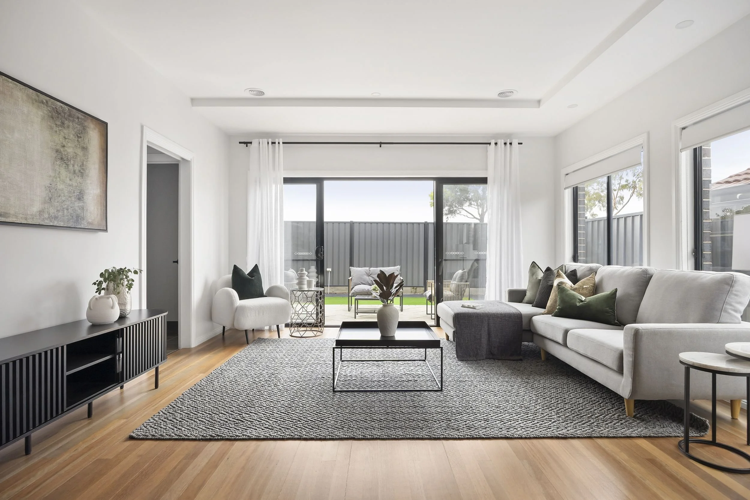 Bright living room with white walls, large windows, and sliding glass door leading outside. Contains a white sofa with green and beige pillows, a gray ottoman, and a black coffee table with a vase of plants. Rug on wooden floor and minimal decor.