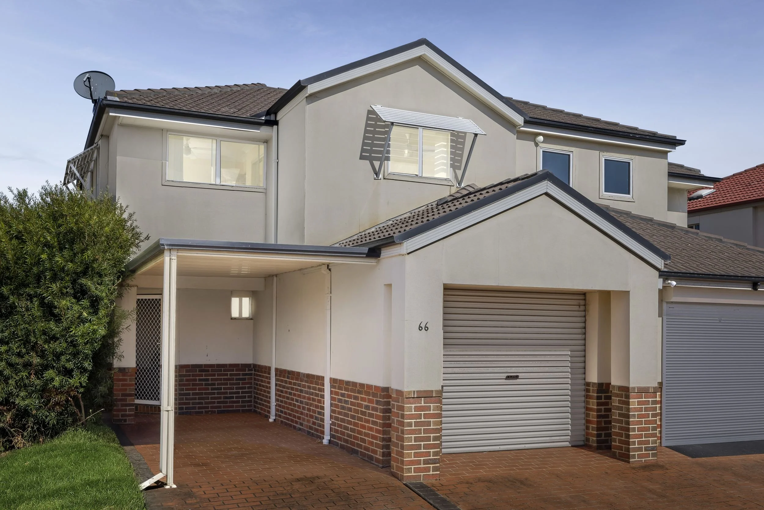 A two-story house with a beige exterior, brown tile roof, and brick lower walls. It has a carport with a brick floor, a garage with a closed roller door, and several windows, including one with an Awnet. There is greenery in the front yard and a satellite dish on the roof.