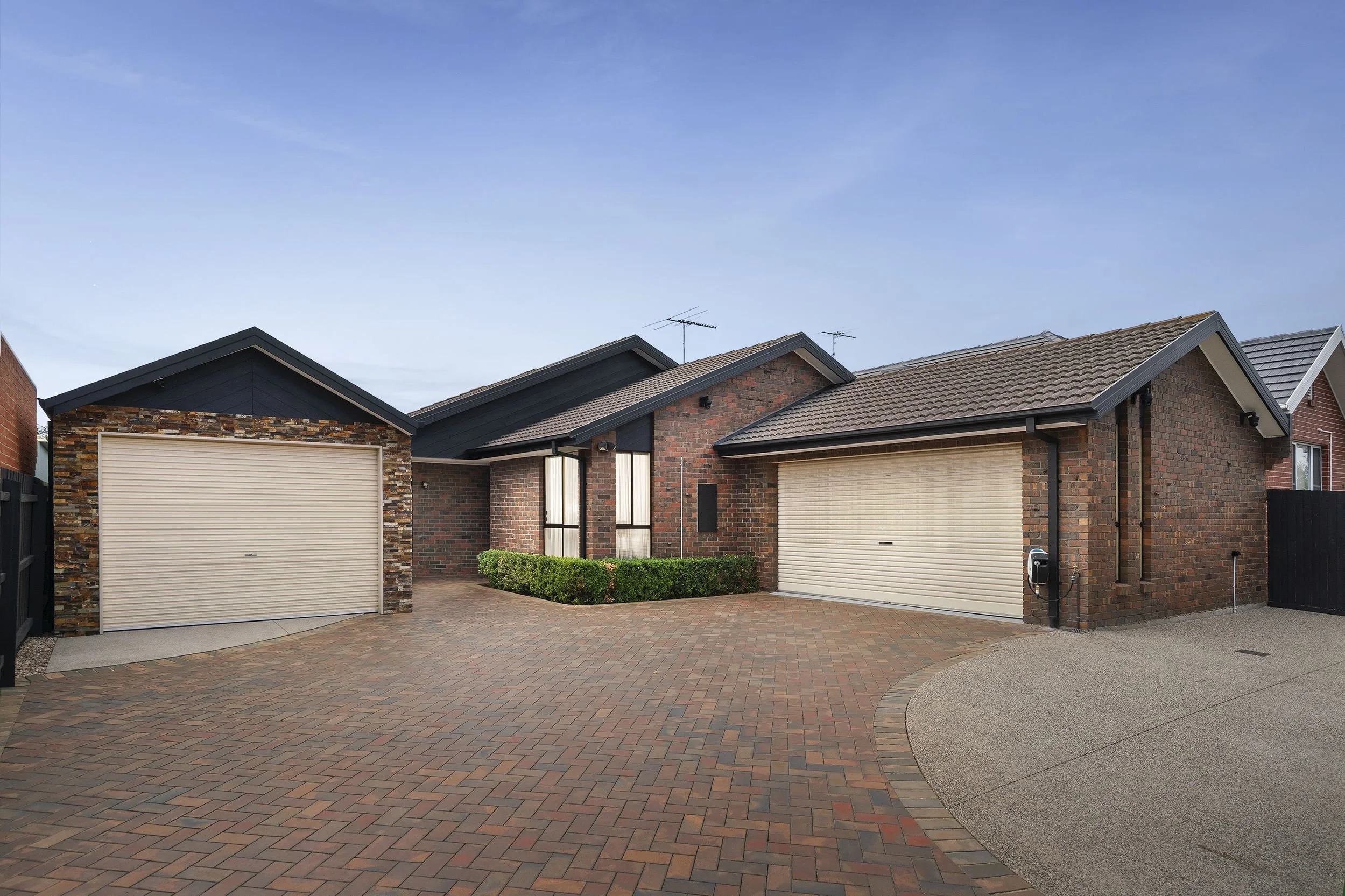 Modern suburban house with two garages, brick and black exterior, curved brick driveway, and a brightly painted well-maintained front yard.