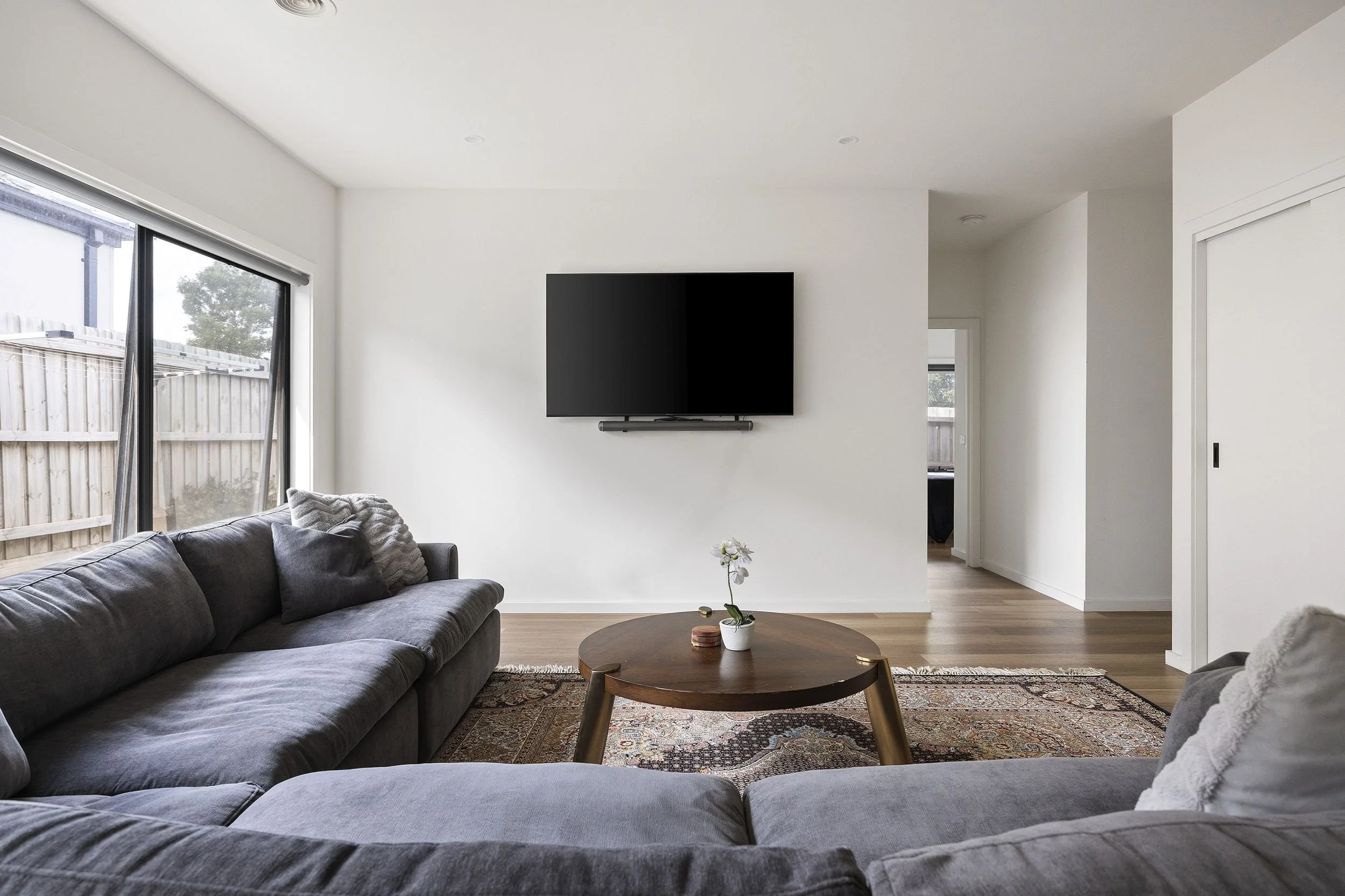 Living room with gray sofa, round wooden coffee table, patterned rug, wall-mounted flat-screen TV, sliding glass door to outdoor space, and white walls.