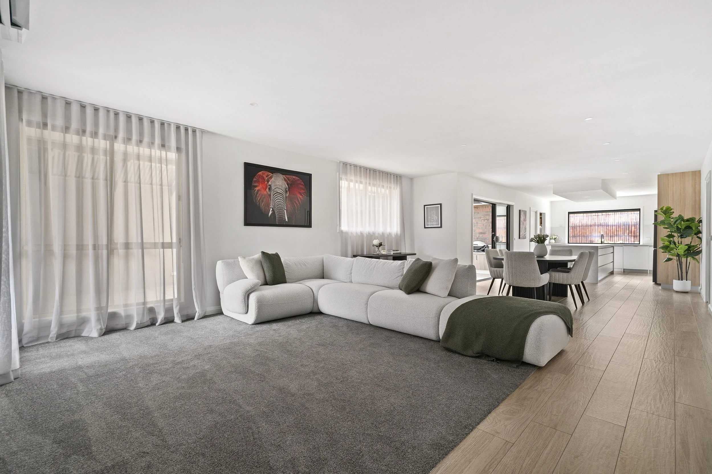 Modern open-concept living room and kitchen with white walls, large windows with sheer curtains, sectional white sofa with green pillows, framed elephant artwork, dining table with six chairs, and kitchen with large window, white cabinetry, and wooden flooring.