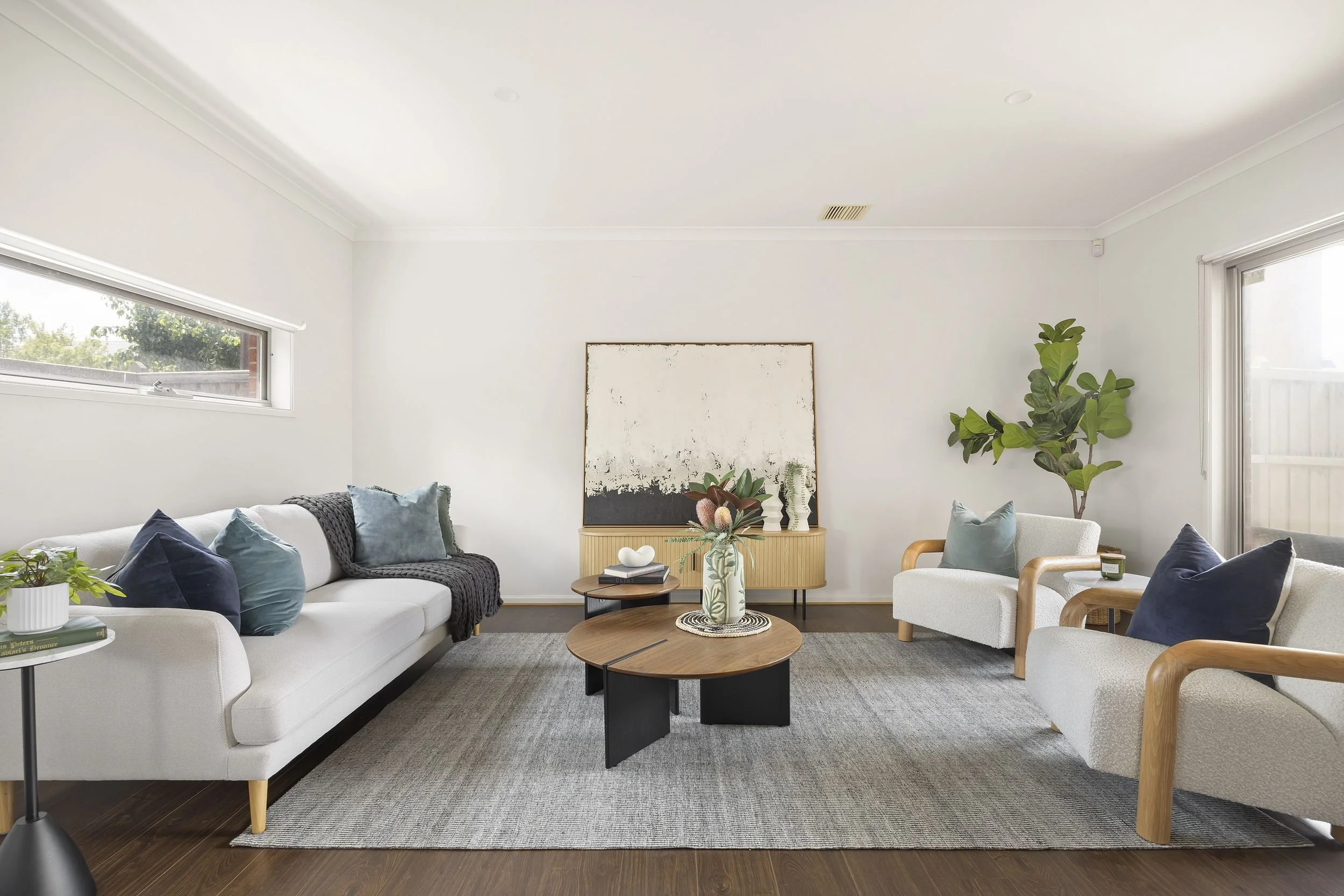 Living room with white walls, large window, and modern furniture including white sofa and armchairs with blue and gray pillows, a wooden coffee table with a vase, a side table with books, a large artwork behind a wooden console, and potted plants.