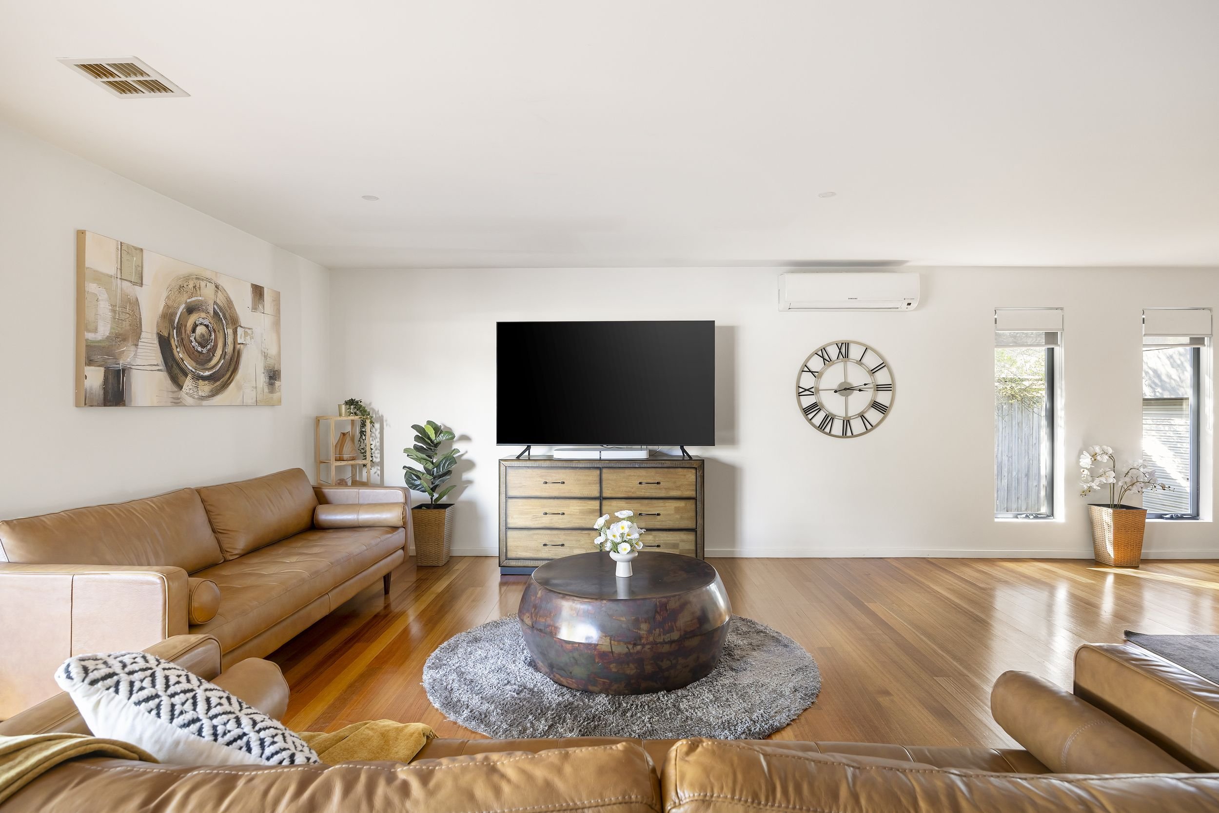 Living room with tan leather sofas, a wooden coffee table with flowers, a large flat-screen TV on a wooden dresser, a decorative wall clock, artwork, indoor plants, and large windows with blinds, hardwood flooring, and a white wall.