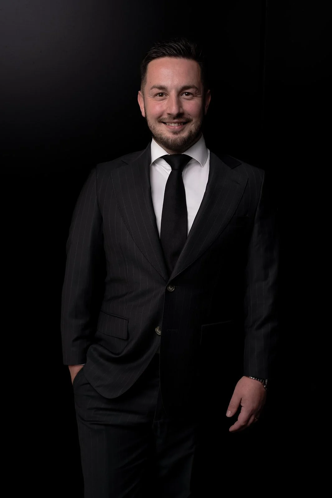 Man in a black suit with a white shirt and black tie, smiling against a dark background.