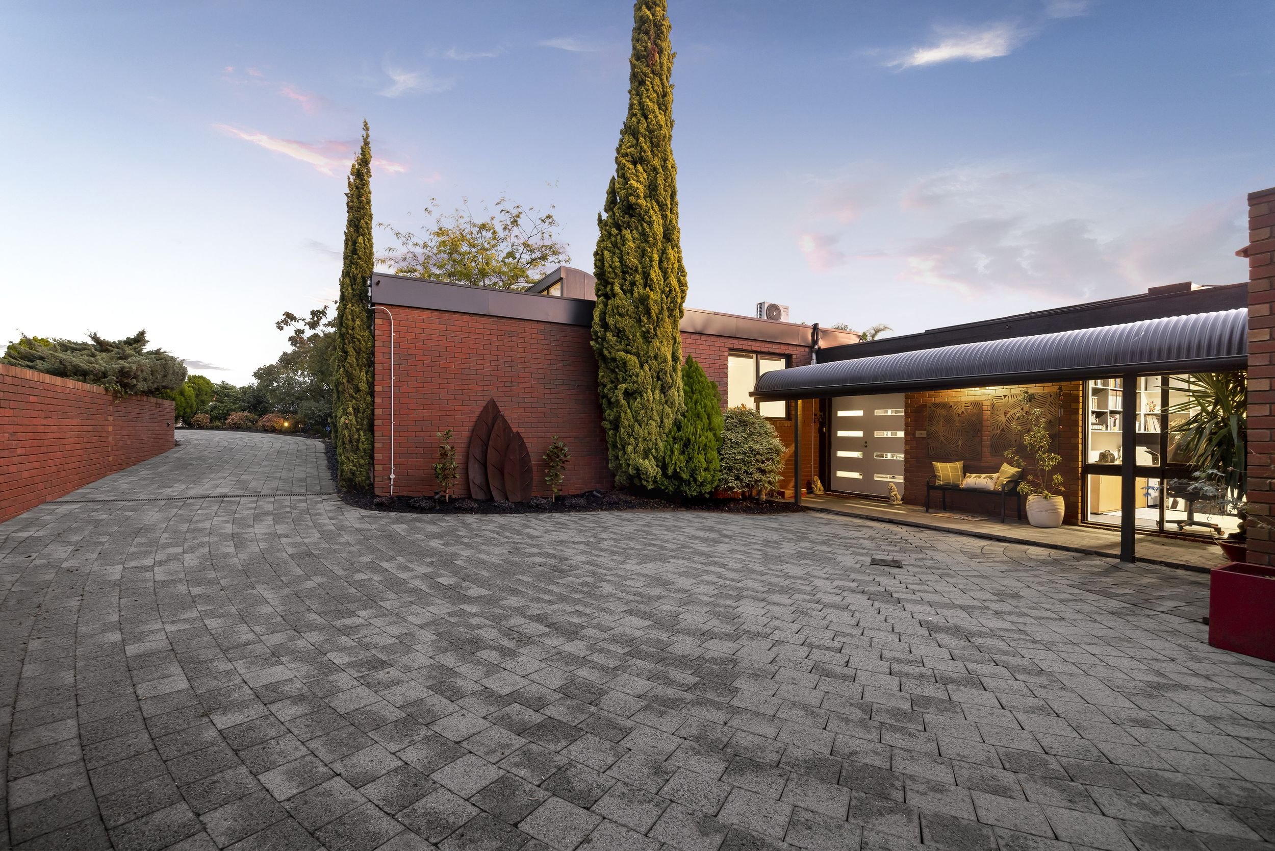 A modern house with a brick exterior, tall trees, a curved stone driveway, and a covered patio with seating at sunset.