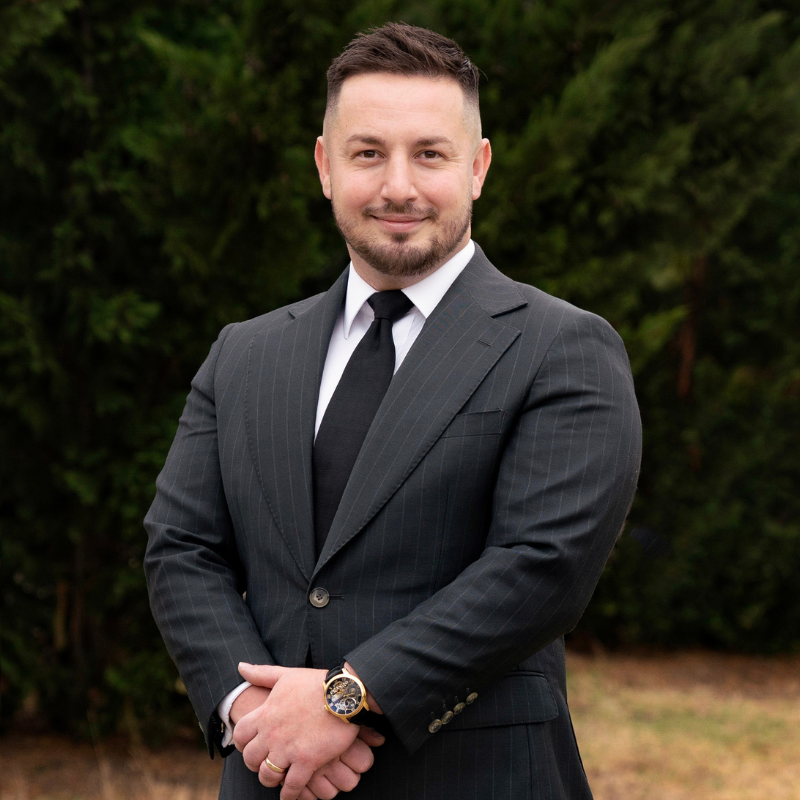 Man in a black suit with a white shirt and black tie, smiling against a dark background.