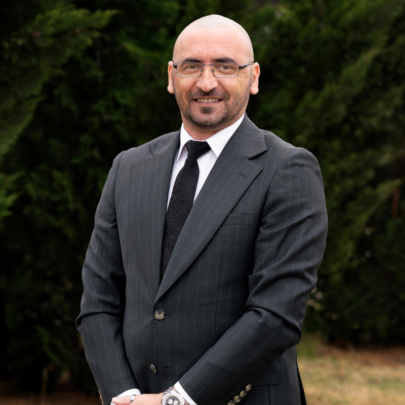 Man in a dark pinstripe suit and glasses against a black background, smiling and posing with hands clasped.