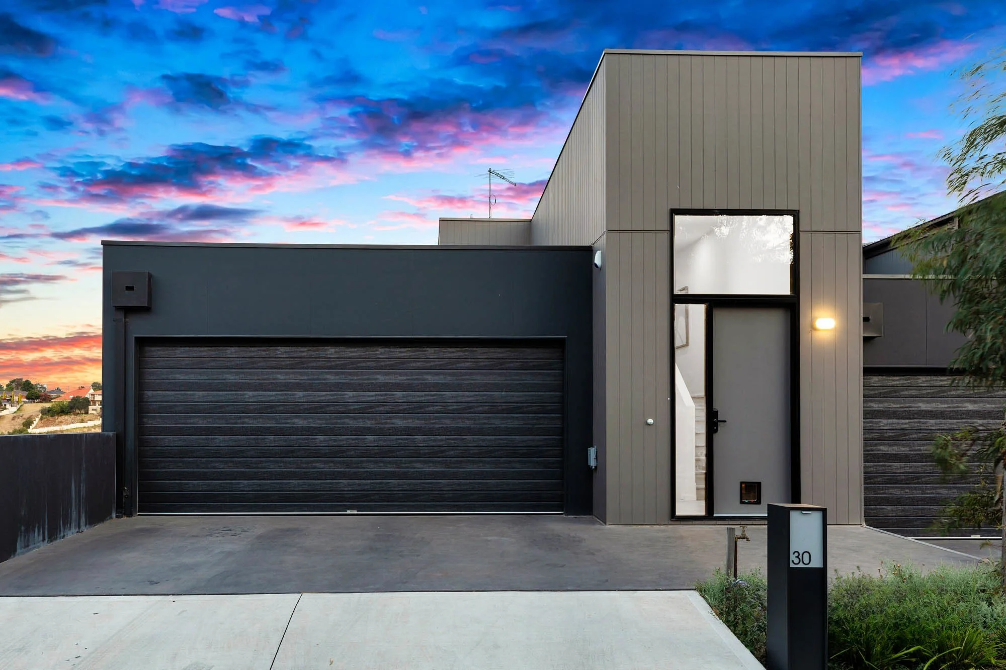 Modern house with black garage door and tall front entrance door, sunset sky with pink and blue clouds, minimal front yard with sidewalk and mailbox.
