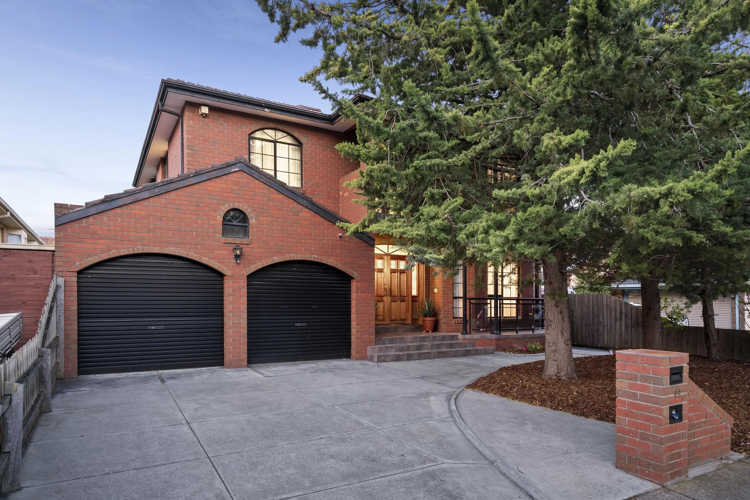 A two-story brick house with a driveway, double garage with black roll-up doors, steps leading to a wooden front door, and a large evergreen tree in a landscaped yard.
