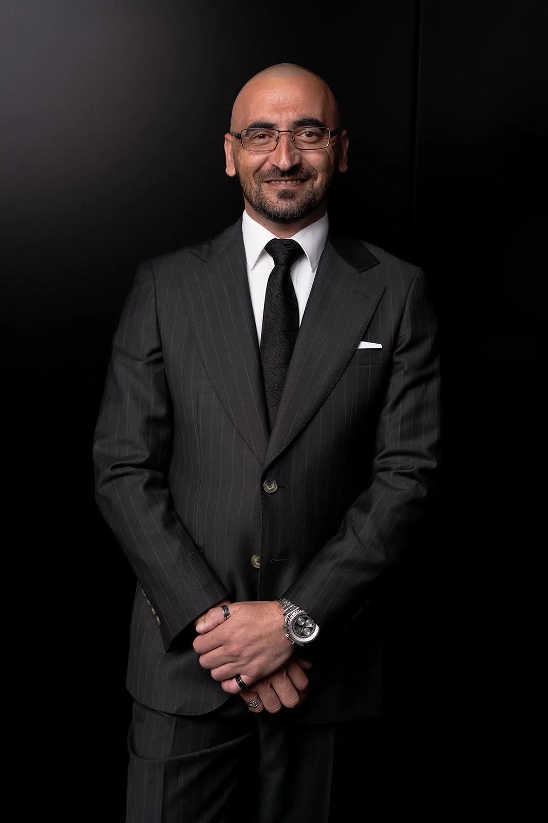 Man in a dark pinstripe suit and glasses against a black background, smiling and posing with hands clasped.