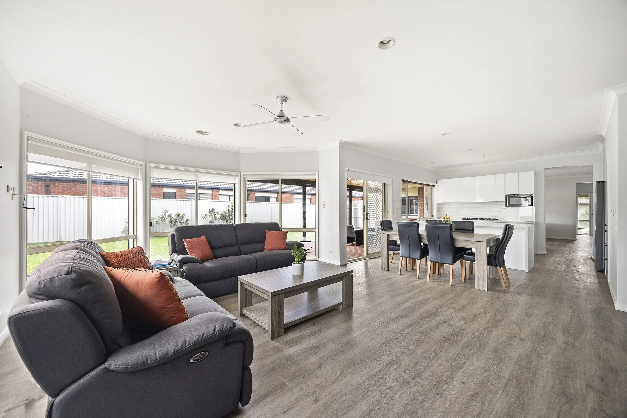 Open-concept living room and kitchen with large windows, gray sofas with rust-colored pillows, a wooden coffee table, a dining table with six black chairs, and white kitchen cabinetry.