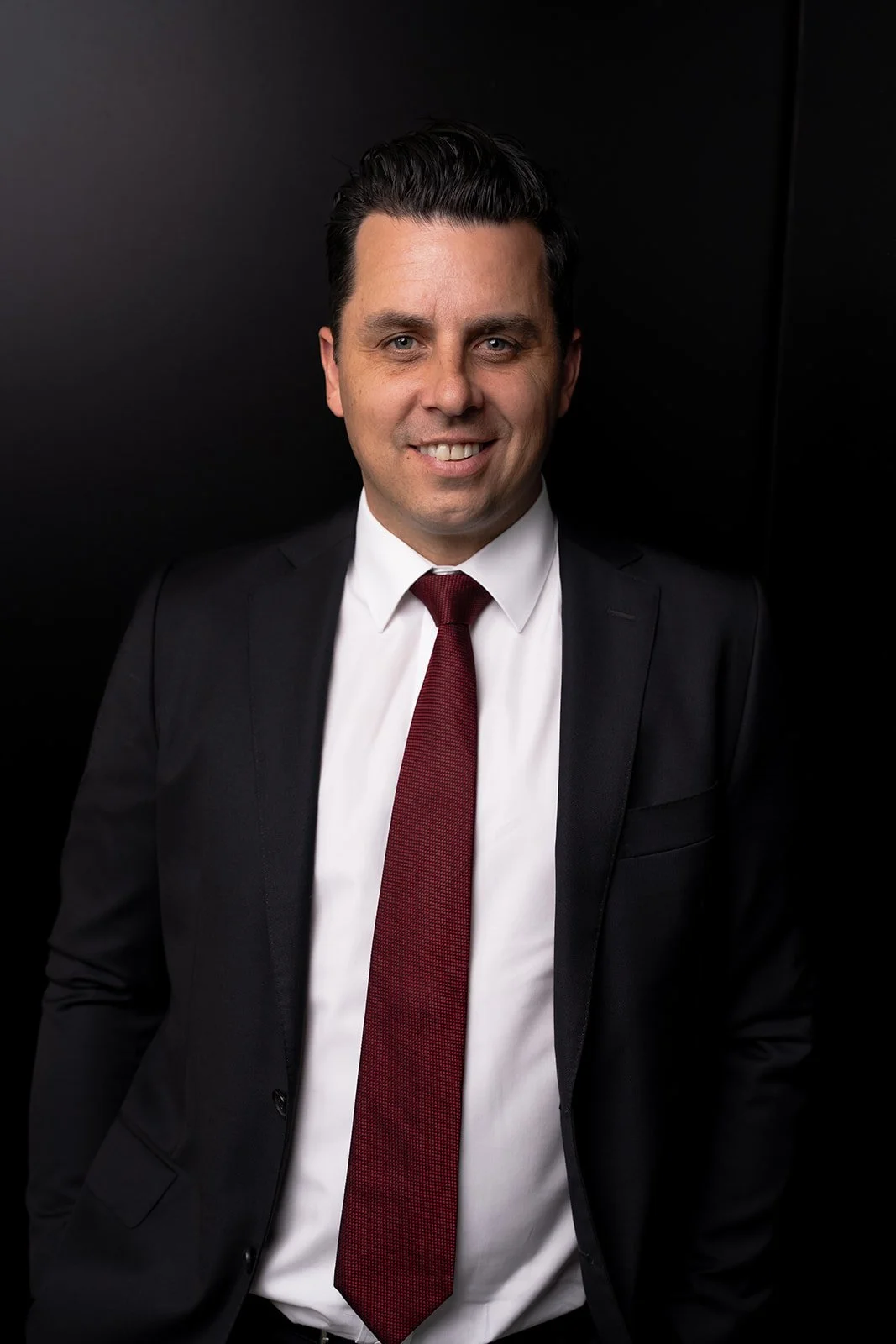 Man in black suit with red tie smiling against black background