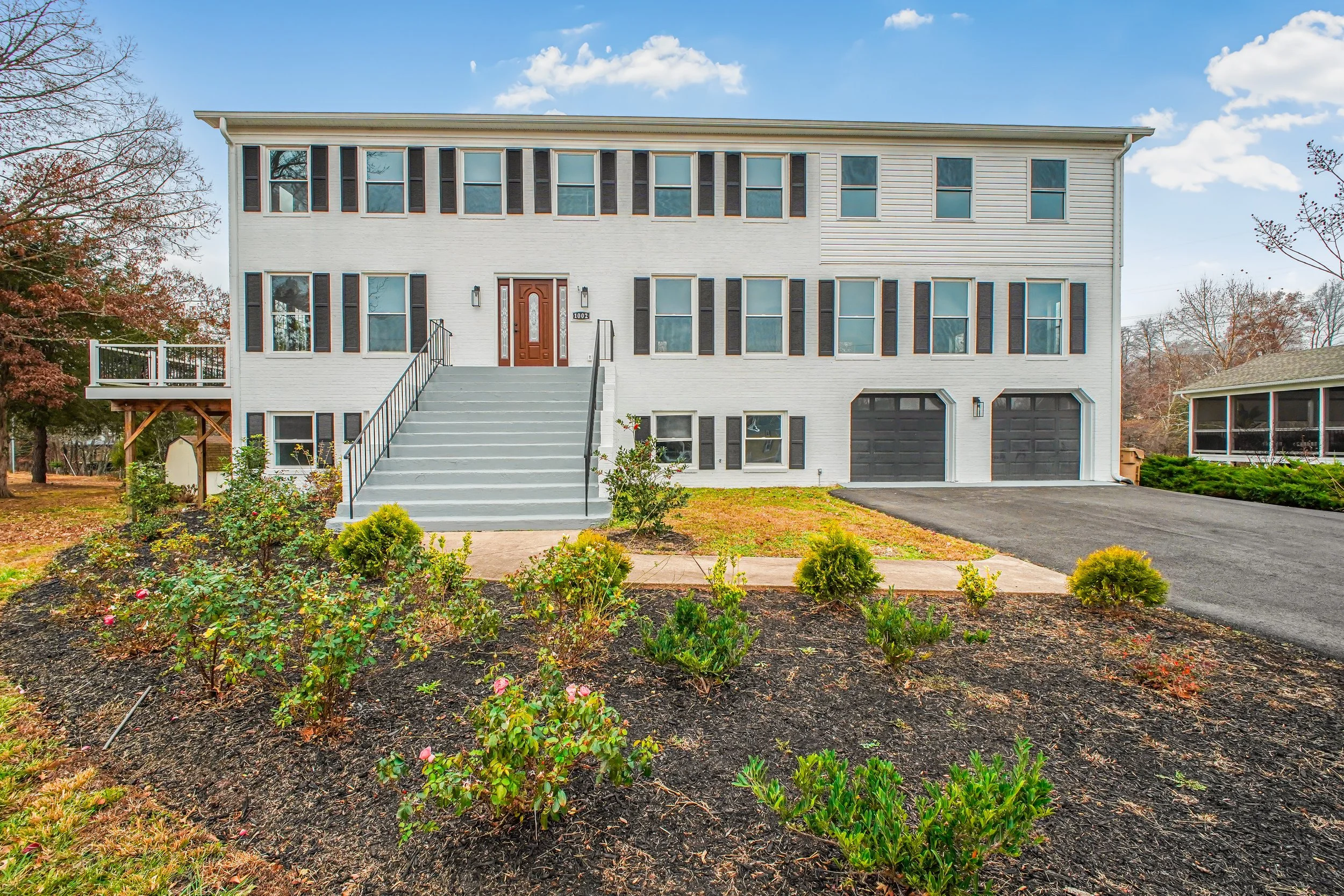 Front view of a multi-story house with a white exterior, black shutters, and a brown front door, with a landscaped yard and driveway.