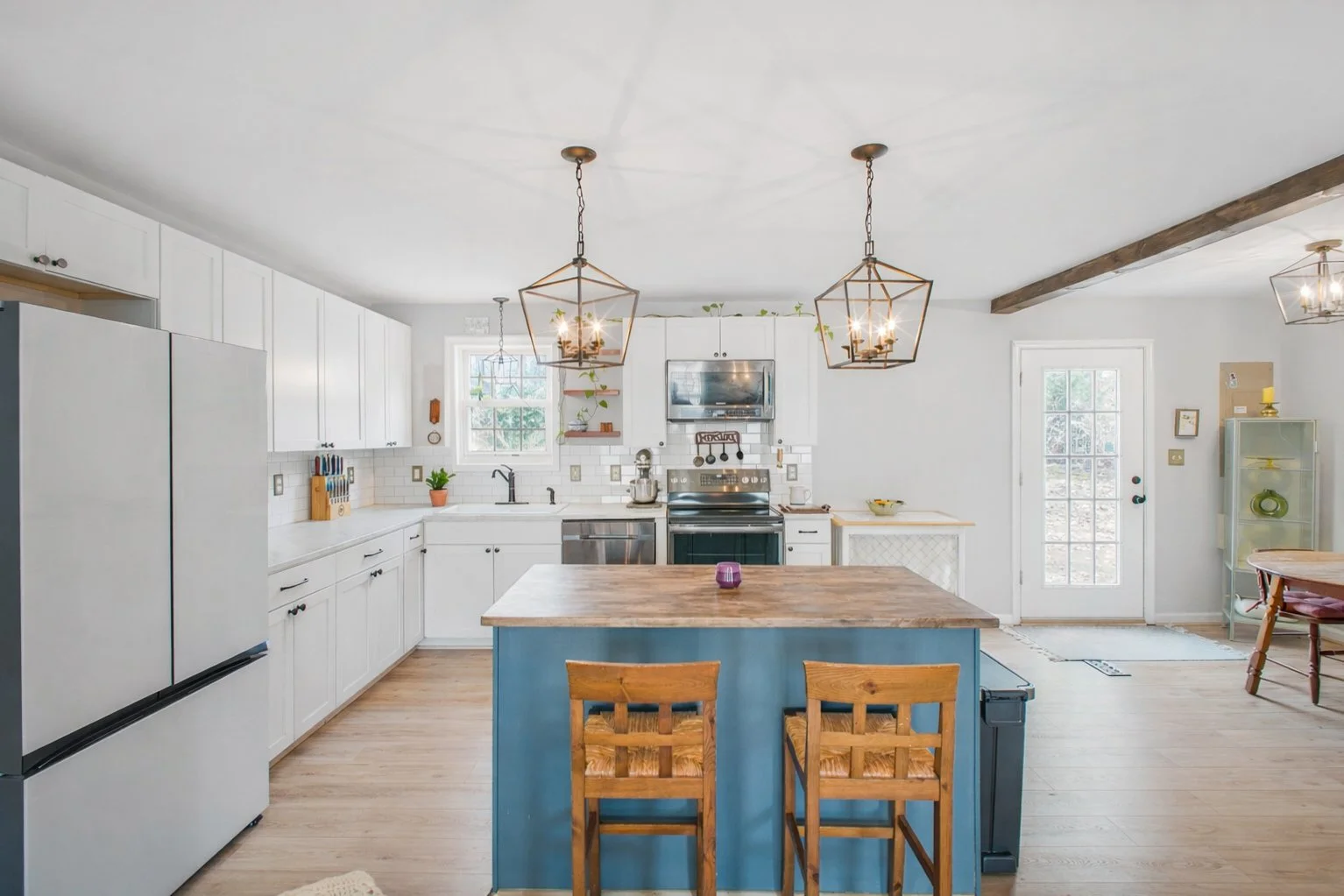 Bright, modern kitchen with white cabinets, a blue island, and wooden chairs, featuring pendant lighting and a wooden beam on the ceiling.