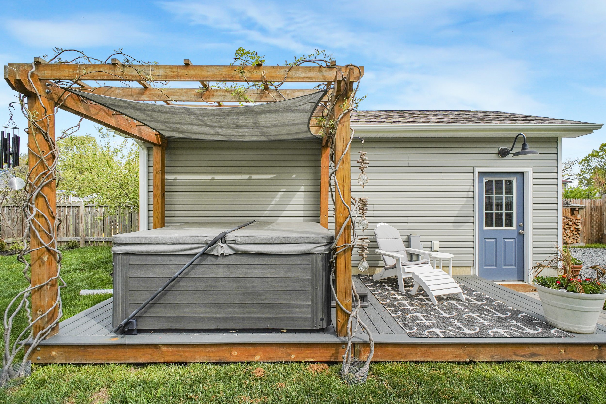 Backyard patio with wooden pergola, hot tub, white Adirondack chair, potted plants, and outdoor rug in front of a house with beige siding and a blue door.