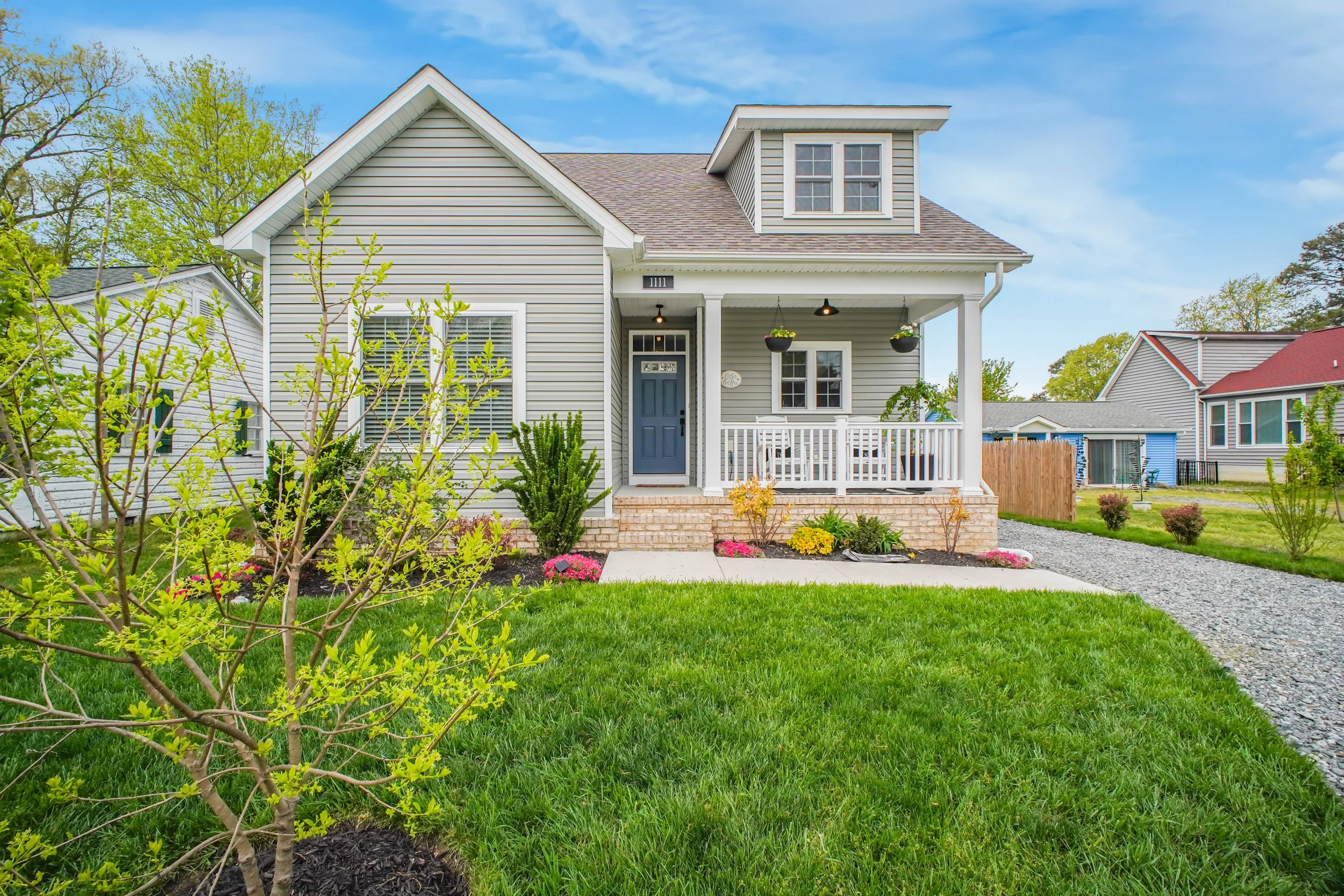 A two-story house with grey siding, a covered front porch, and a gabled roof, surrounded by a lush green lawn and blooming flowers.