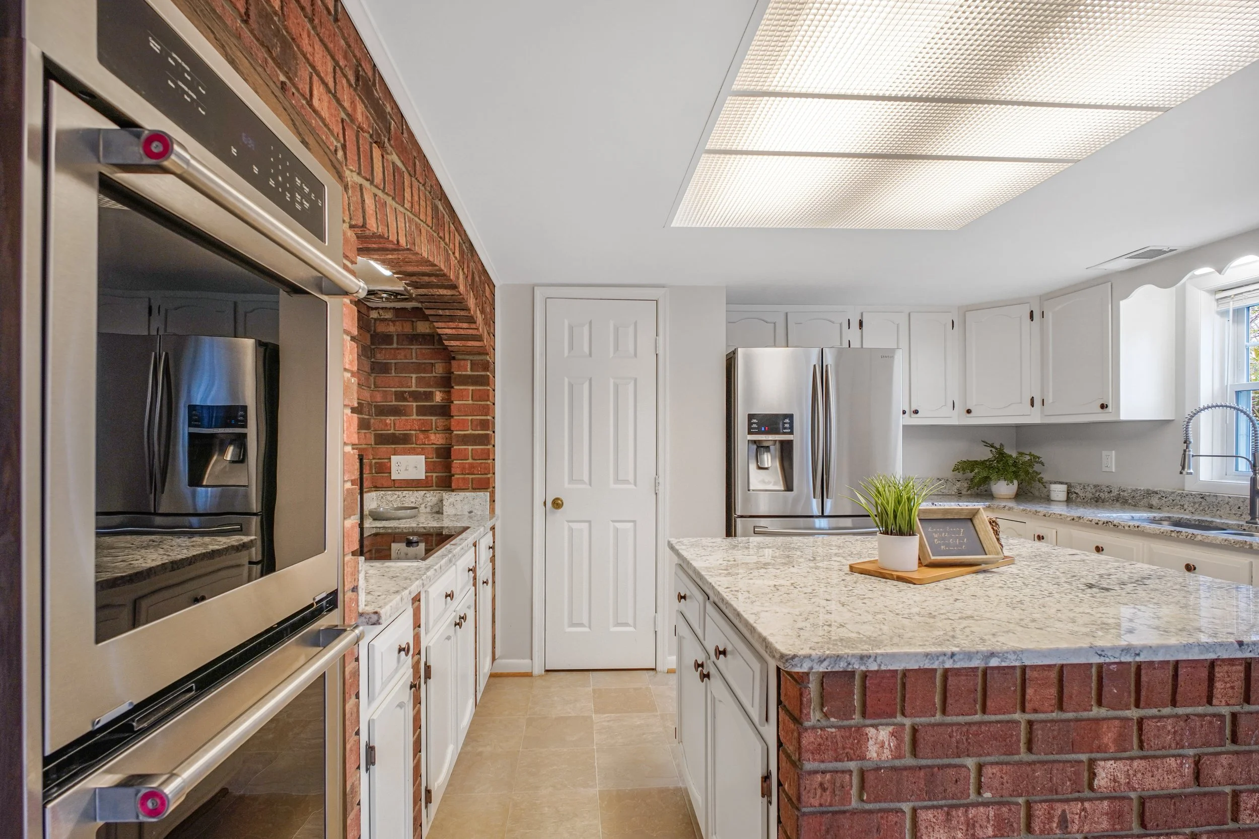 A modern kitchen with white cabinets, granite countertops, built-in stainless steel appliances, a red brick arch, and a ceiling with fluorescent lighting panels.