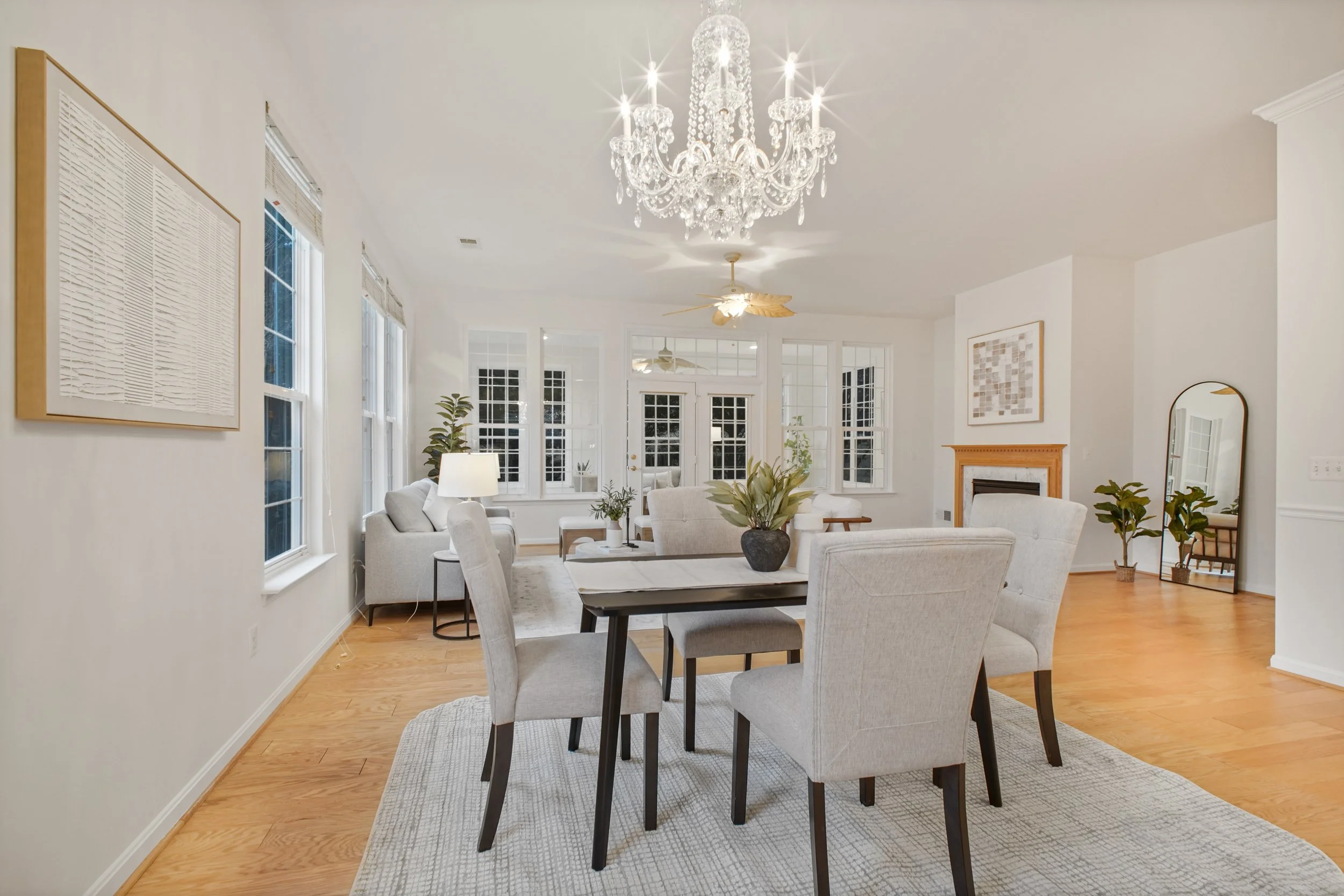 Bright living room with white walls, large windows, chandelier, ceiling fans, white couches, armchairs, coffee table, plants, and artwork.