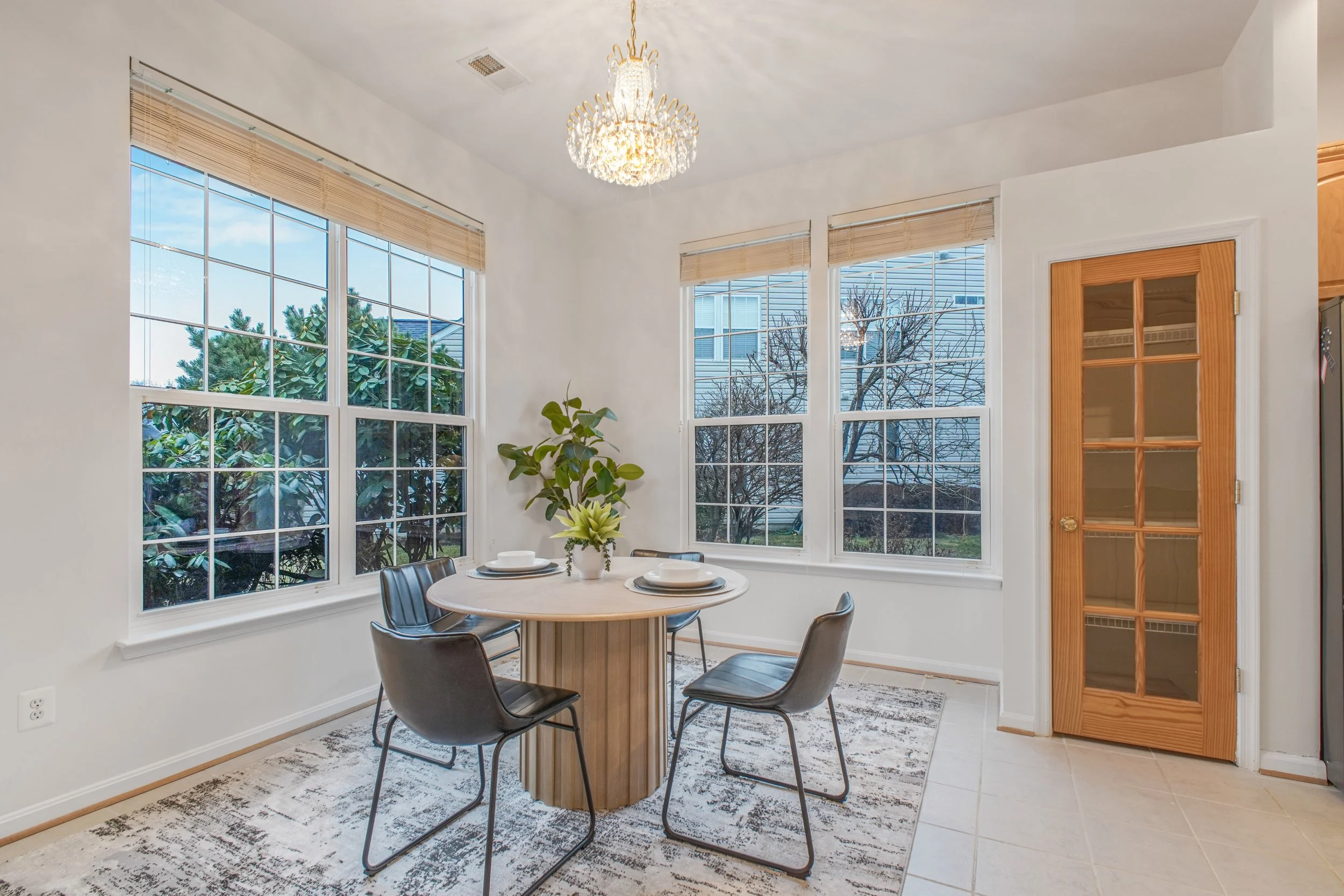 A dining area with a round table set with four plates and a plant in the center, surrounded by five black chairs, large windows with bamboo blinds letting in natural light, a chandelier overhead, a patterned rug beneath the table, and a wooden door on the right leading to another room.