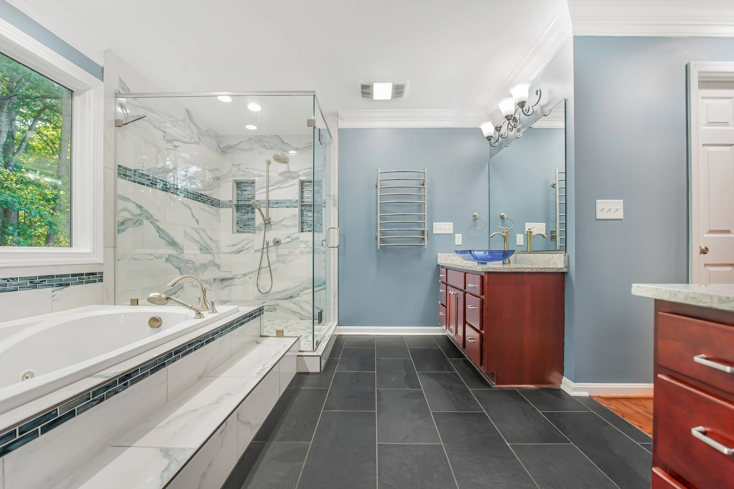 A modern bathroom with a large window showing greenery outside. It has a bathtub with a marble surround, a glass-enclosed walk-in shower with marble and tile accents, a vanity with a blue vessel sink, a red wooden cabinet, a mirror, and wall-mounted light fixtures.