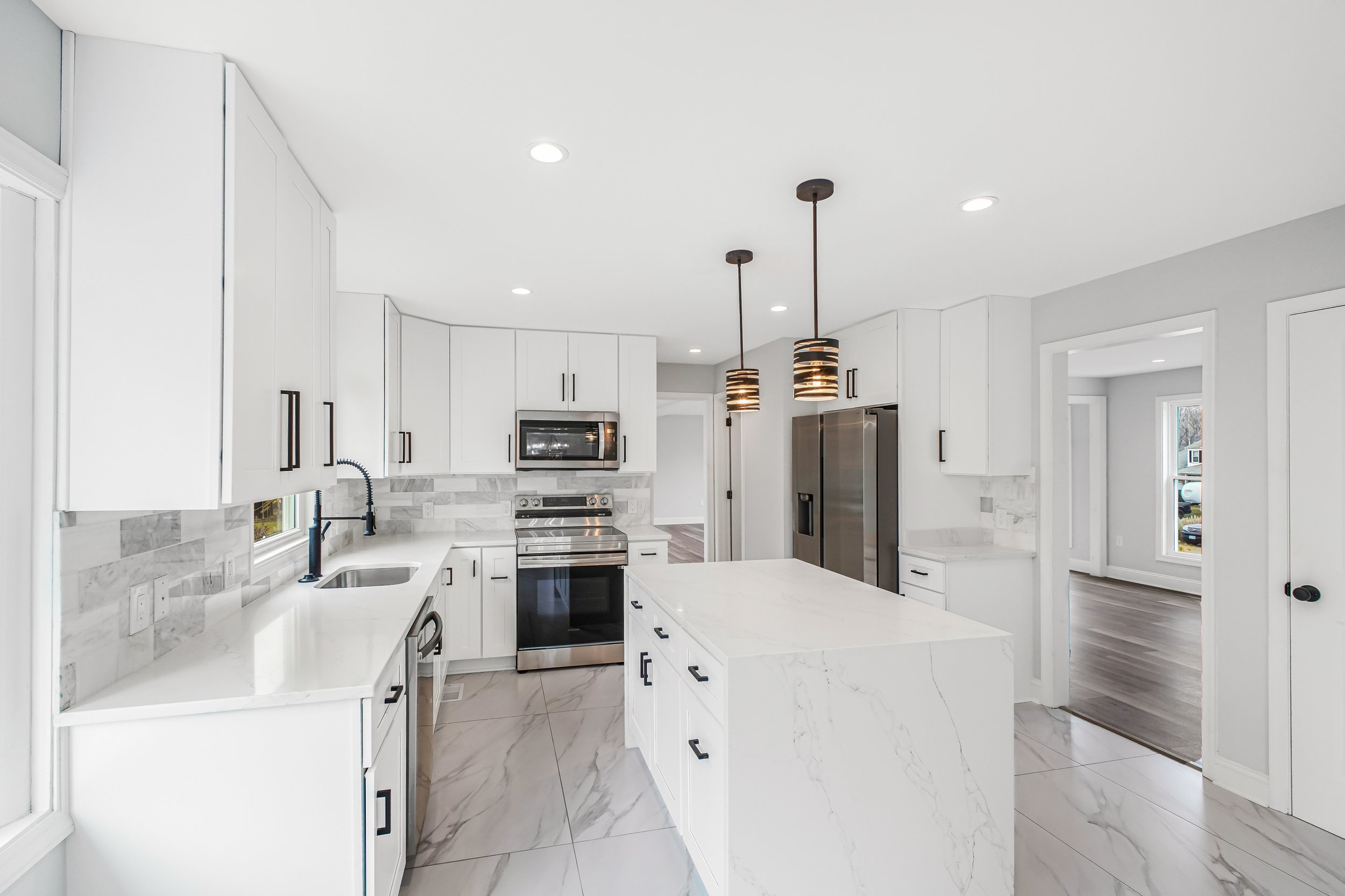 Modern kitchen with white cabinets, marble backsplash, stainless steel appliances, black hardware, pendant lights, and large windows.
