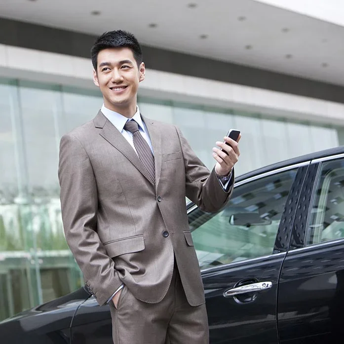 A smiling man in a brown suit holding a phone standing beside a black car in front of a modern building with glass windows.