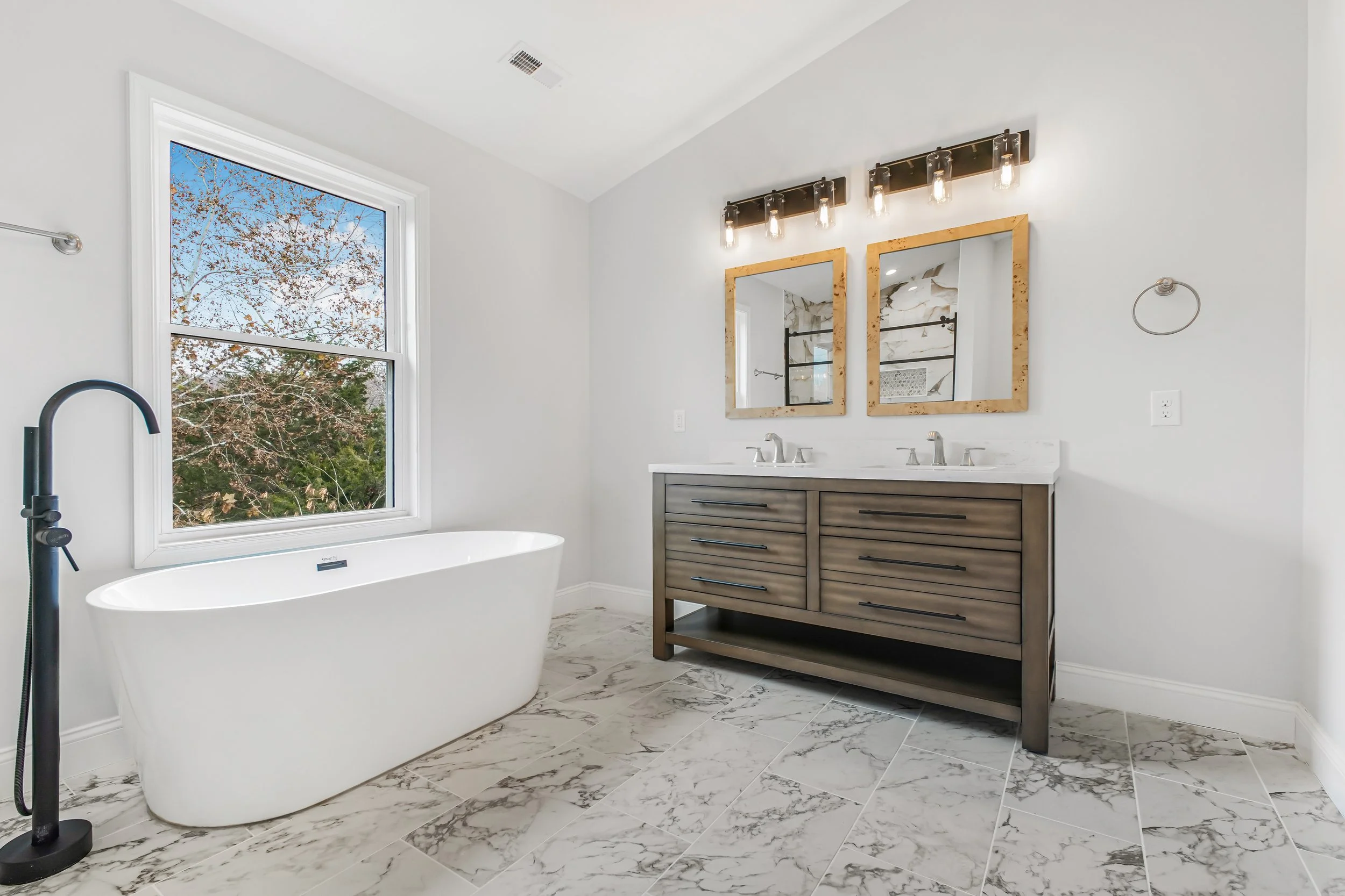 Modern bathroom with a freestanding bathtub near a window, double sink vanity with mirrors, and marble floor tiles.