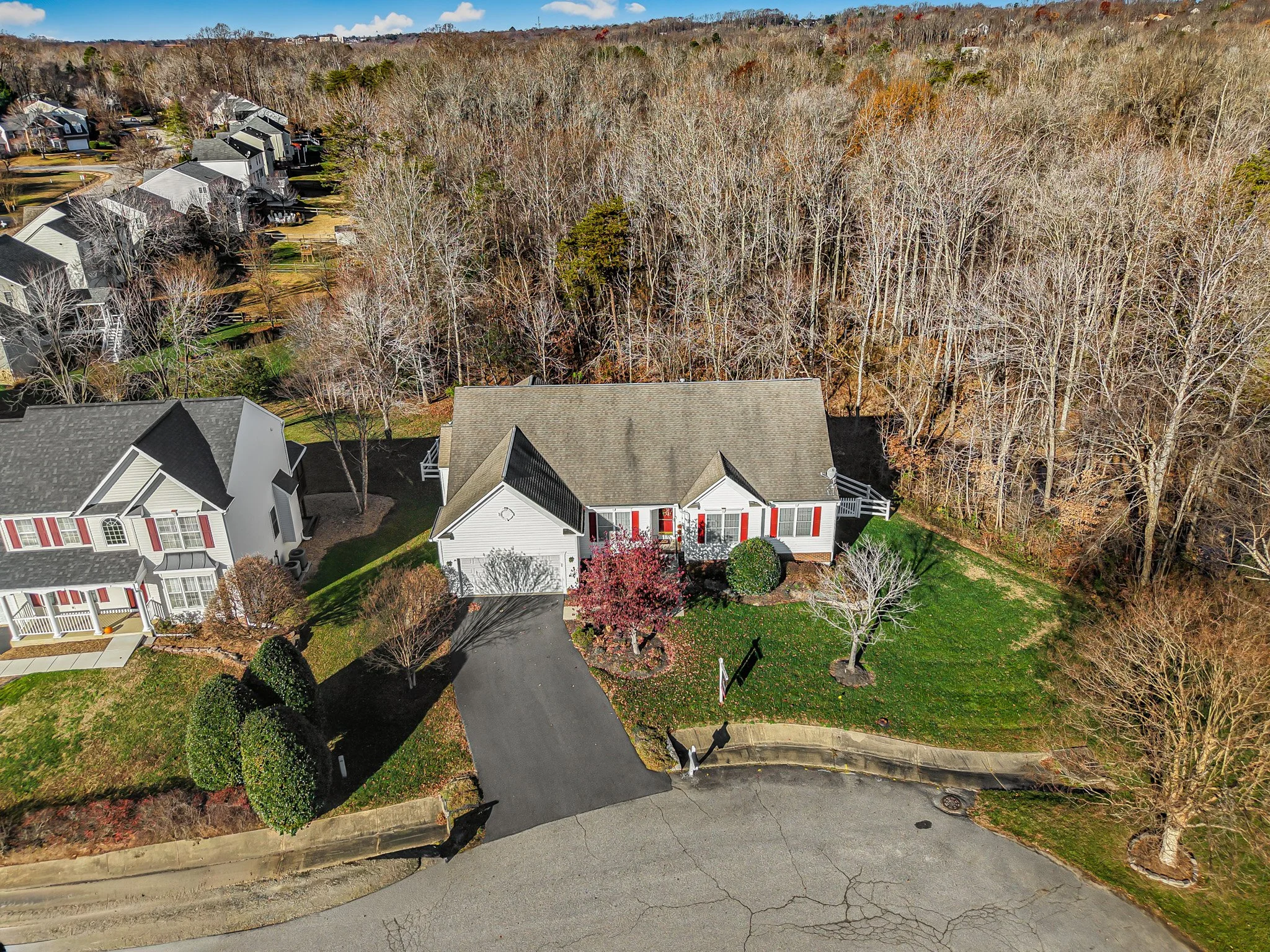 An aerial view of a residential neighborhood in autumn. The image features a white house with red shutters and a gray roof, a driveway, and surrounding trees with no leaves. Adjacent houses with similar architecture are visible, and a wooded area is behind the houses.