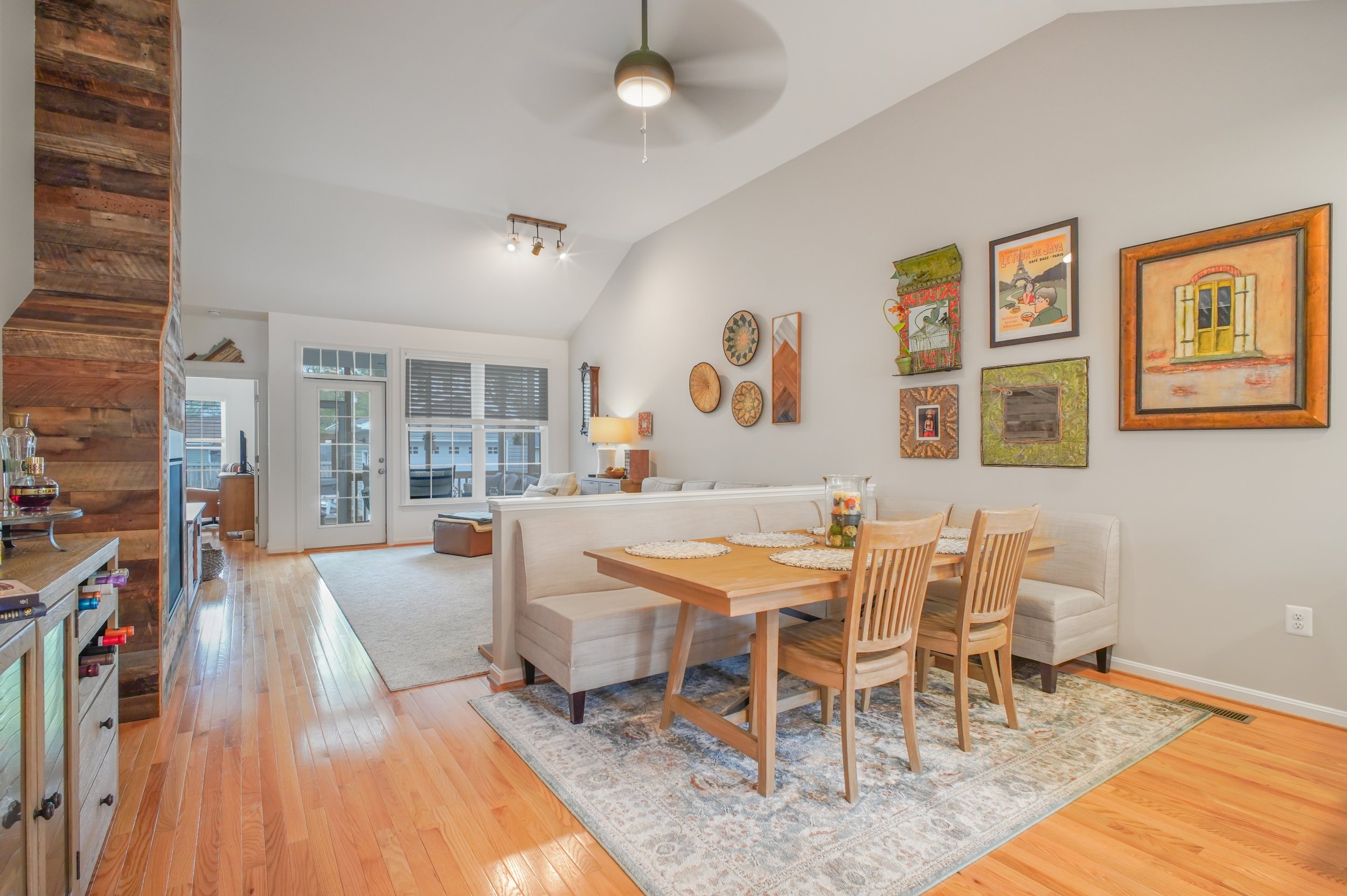 Interior of a living and dining area with wooden flooring, white walls, a wooden dining table with chairs, a patterned area rug, wall art, a white sofa, a side table, and large windows with blinds, illuminated by ceiling and wall lights.