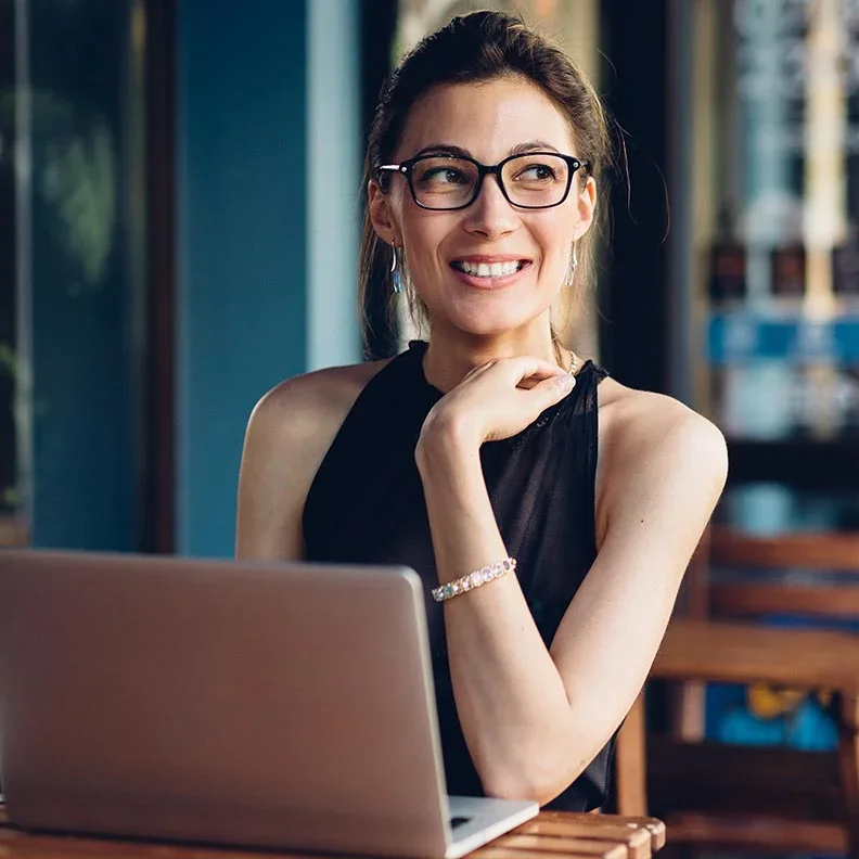 A woman with glasses, smiling and looking to her right, sitting at a table with a laptop.
