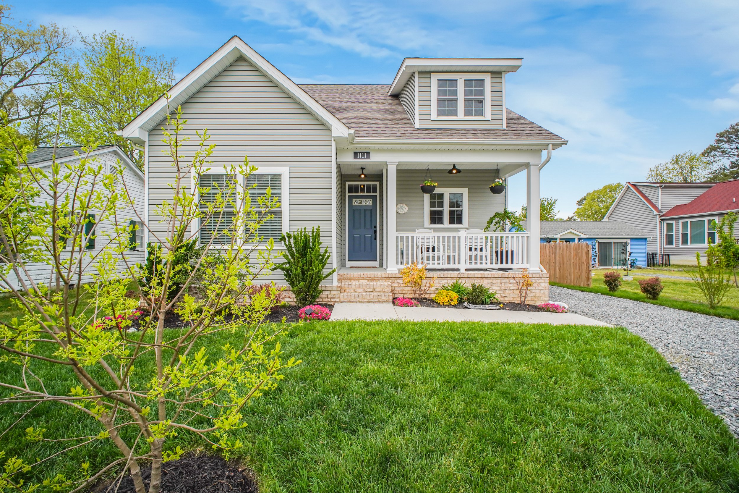 A two-story house with grey siding, white trim, and a front porch with white railing. There are hanging flower baskets, a blue front door, and well-maintained garden beds with colorful flowers in the yard. The lawn is green, and a gravel pathway leads to the porch. The background includes neighboring houses and trees during springtime.