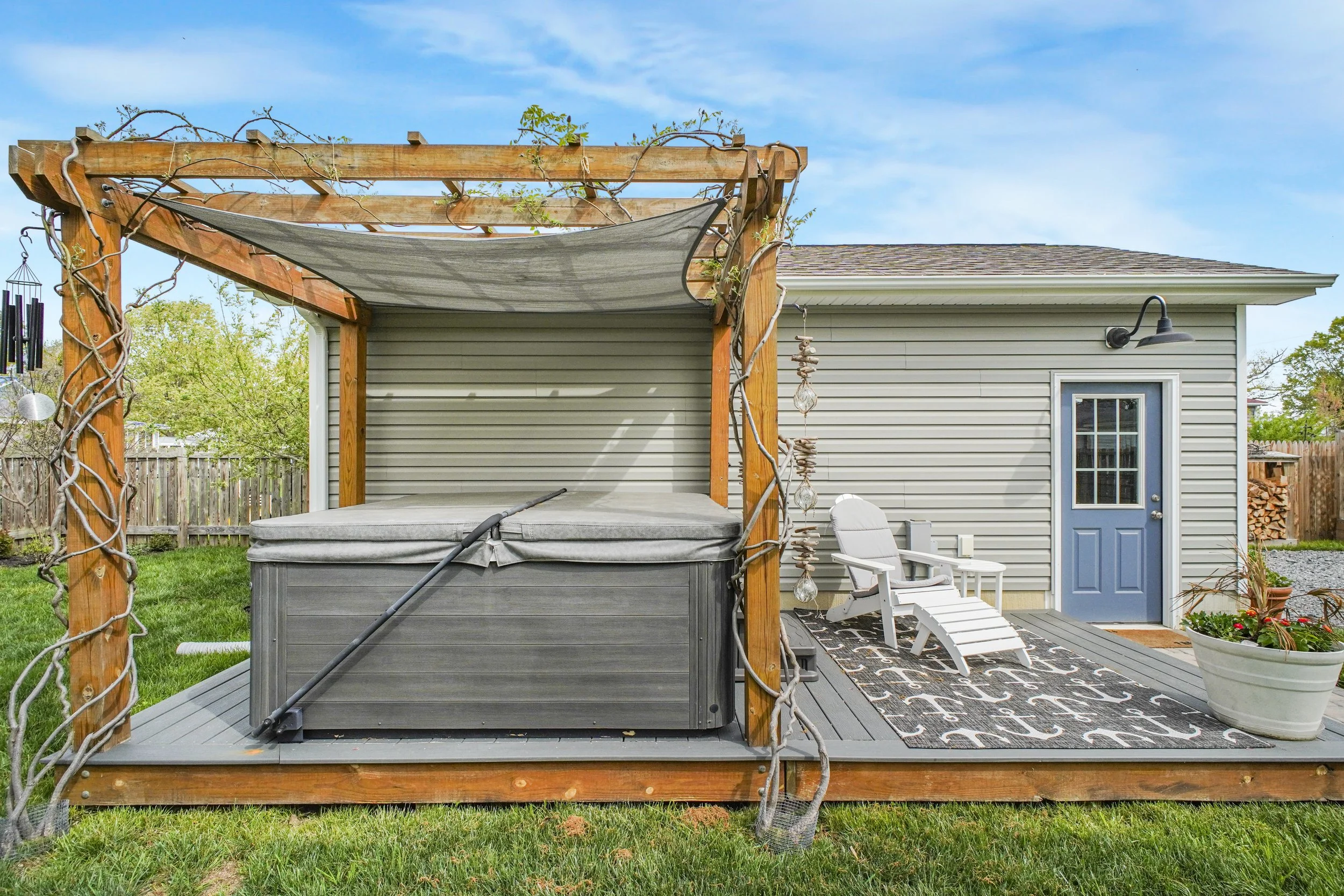 Backyard patio with a pergola covered with vine, hot tub, outdoor table and chairs, door to house, and decorative rug under the seating area.