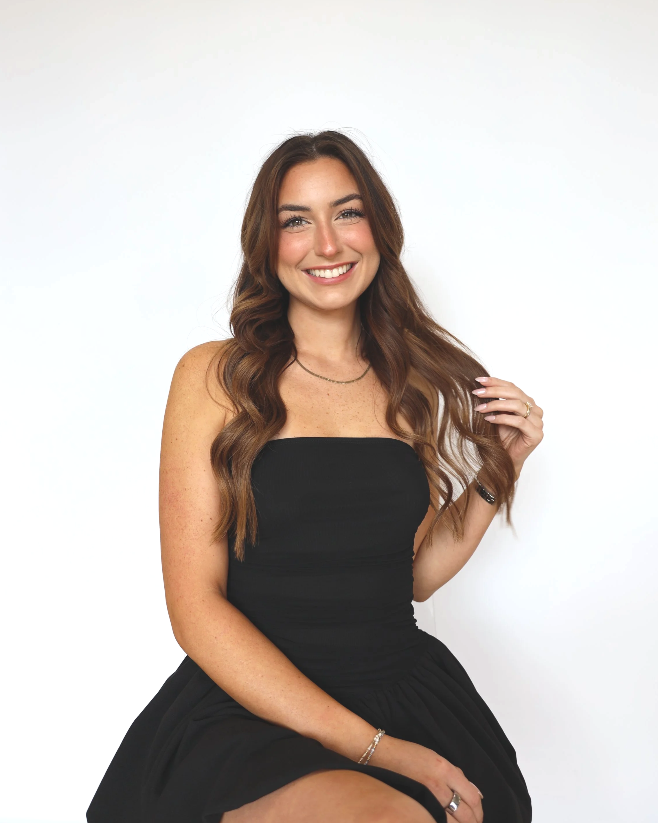 A woman with long wavy brown hair, wearing a black strapless top and a silver necklace, smiling and sitting against a plain white background.