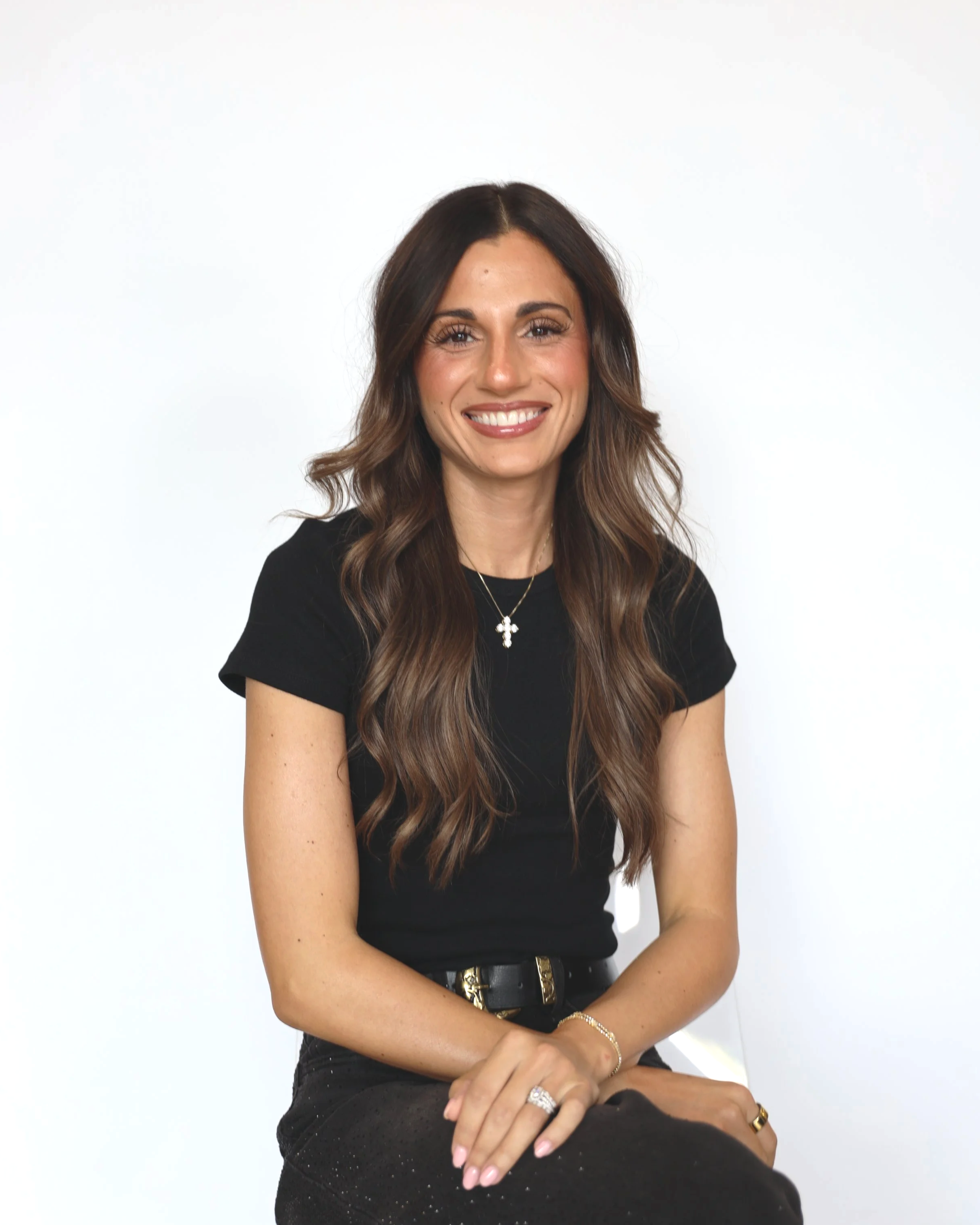 A woman with long wavy brown hair sitting against a plain white background, smiling and wearing a black top, a black belt with gold details, and jewelry including rings, a necklace, and bracelets.