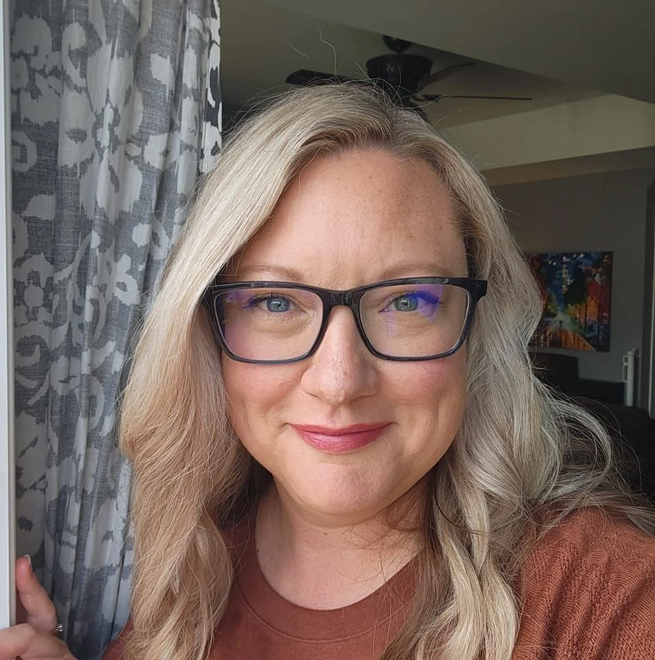 A woman with blonde hair and glasses smiling at the camera indoors.