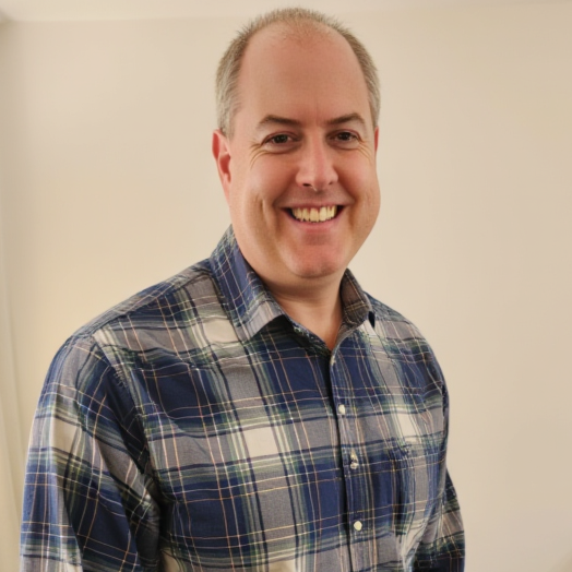 Smiling man wearing a plaid shirt standing in front of a plain wall.