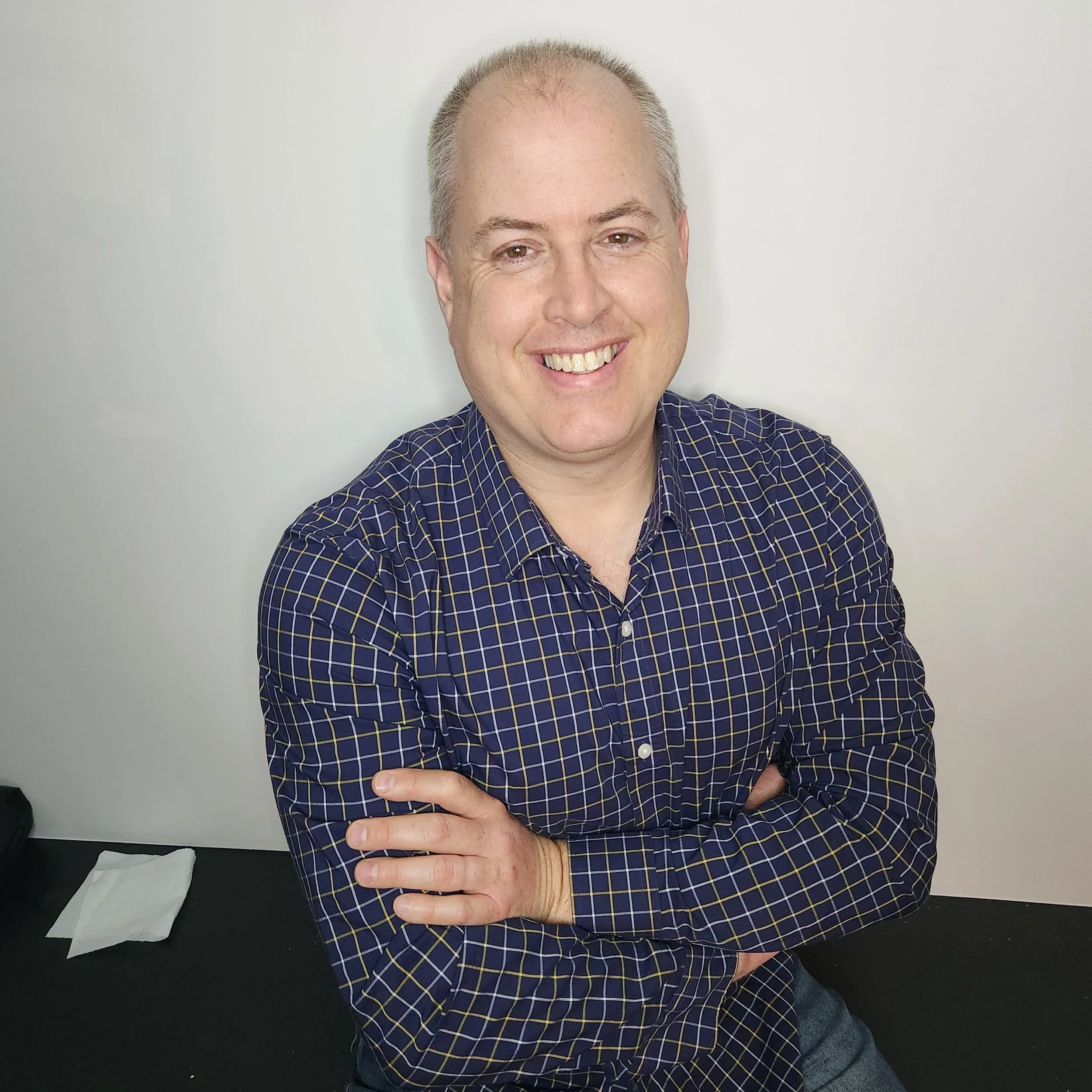 A smiling man with light skin and short gray hair wearing a navy blue checkered shirt, standing with arms crossed against a plain white background.
