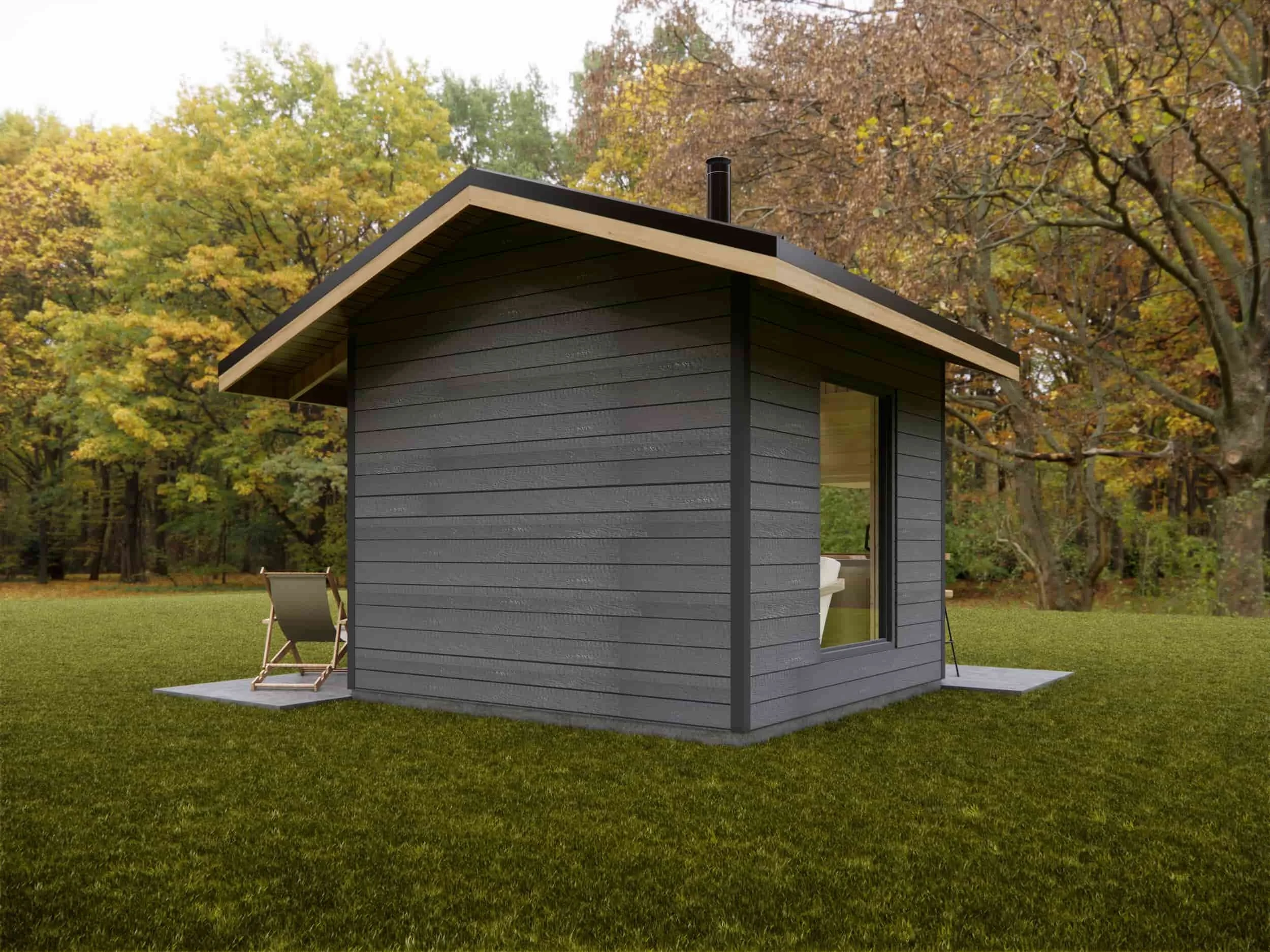 A small modern gray house with a sloped roof, glass sliding door, and two deck chairs outside, surrounded by a grassy yard and trees with fall foliage.