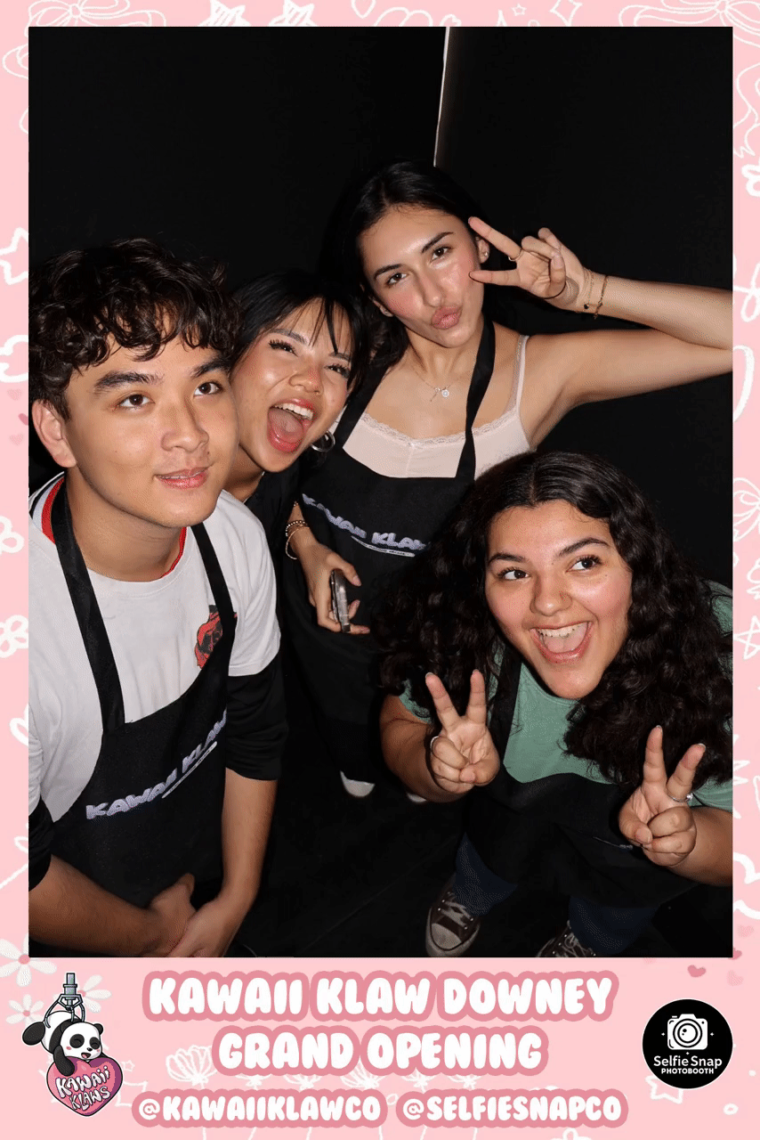 Four people wearing black aprons posing happily in a photo booth at the Kawaii Klaw Downey grand opening. The image has a pink, decorated border with event branding.