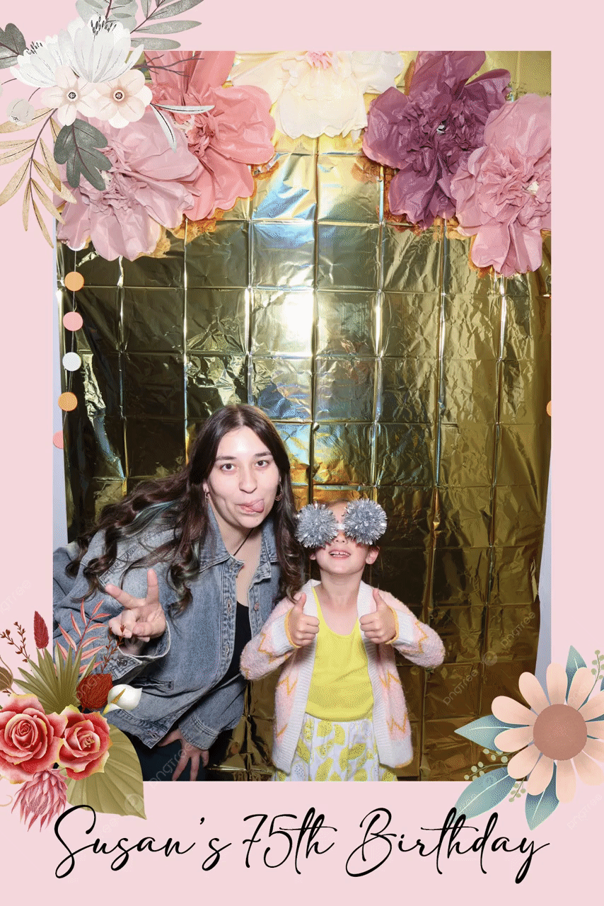 Two people posing in front of a gold backdrop with pink floral decorations. One is sticking out their tongue, the other wearing decorative glasses and giving thumbs up, for Susan's 75th Birthday celebration.
