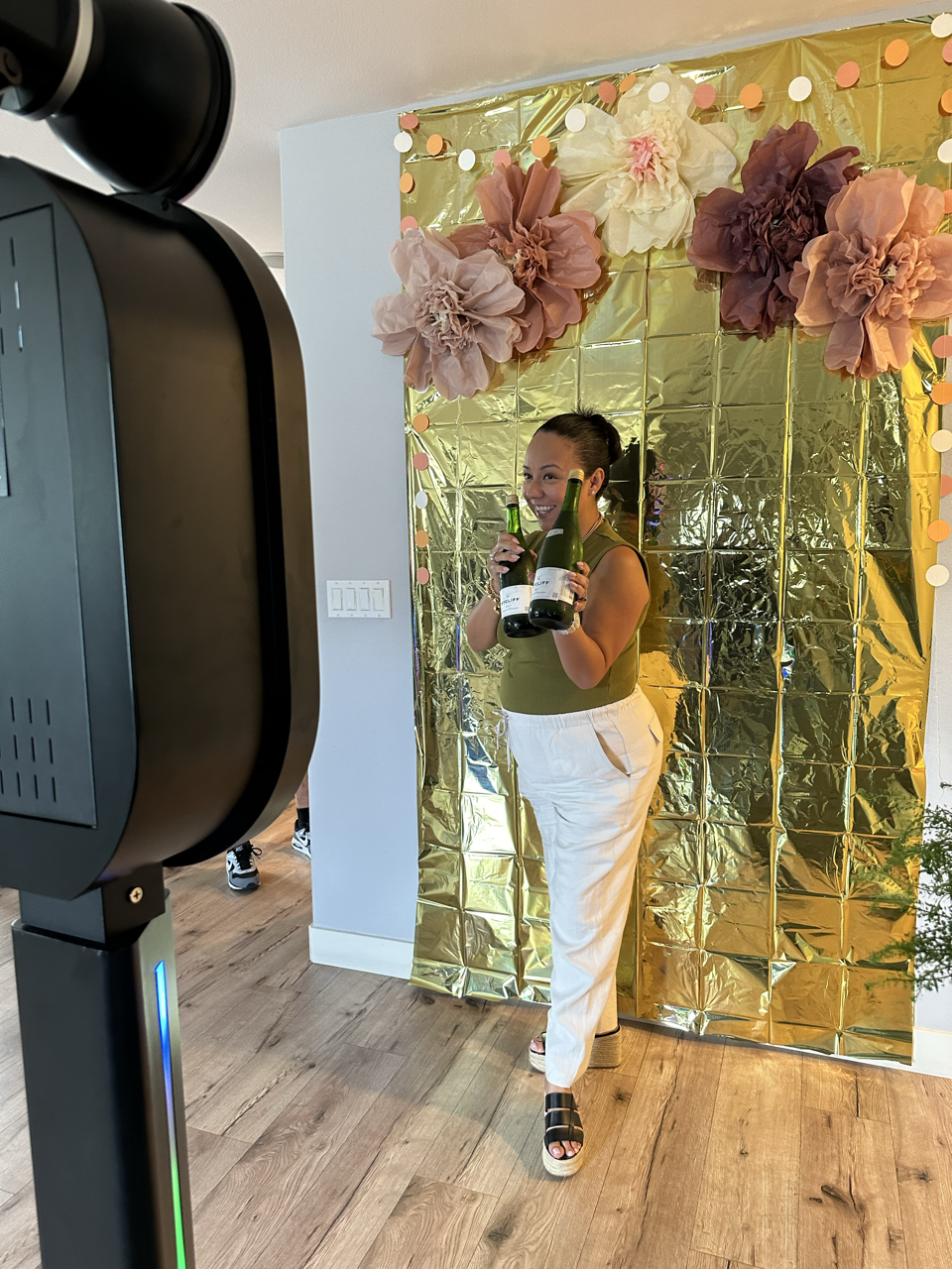 Person posing for a photo booth with gold foil backdrop and large paper flowers, holding two champagne bottles, wearing a green top and white pants.