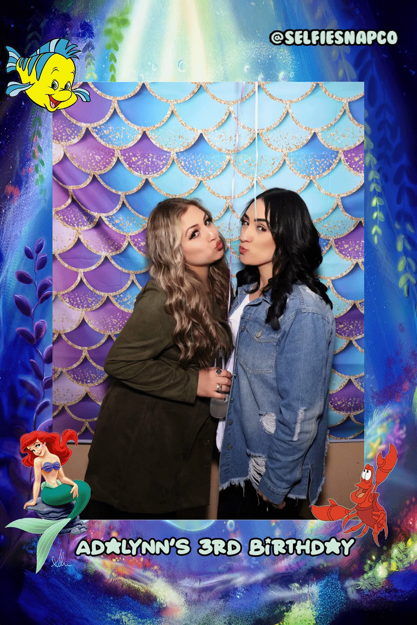 Two women posing for a photo in front of a mermaid-themed backdrop with "Adalynn's 3rd Birthday" text, featuring cartoon sea characters. They are making playful expressions.