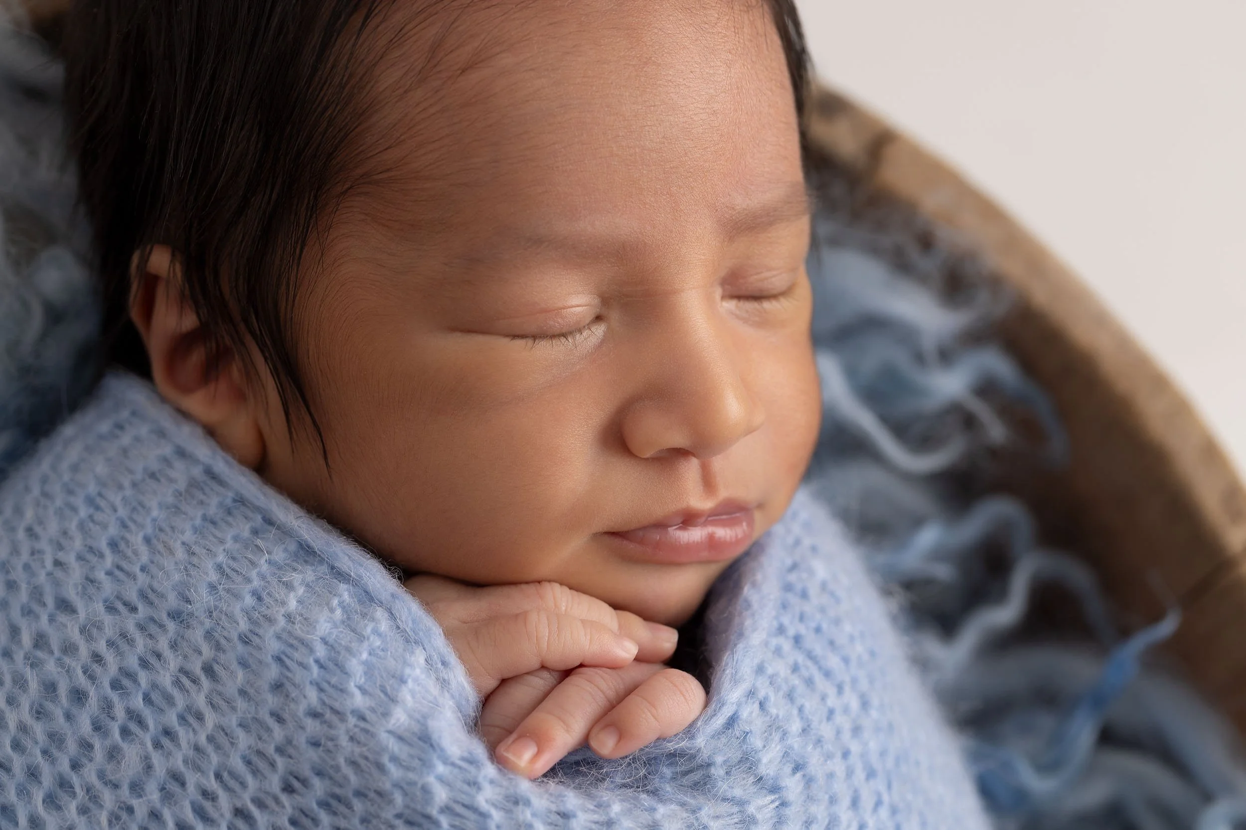 Close-up of a sleeping newborn baby with closed eyes, peacefully resting with hands tucked under chin, wrapped in a soft blue blanket.