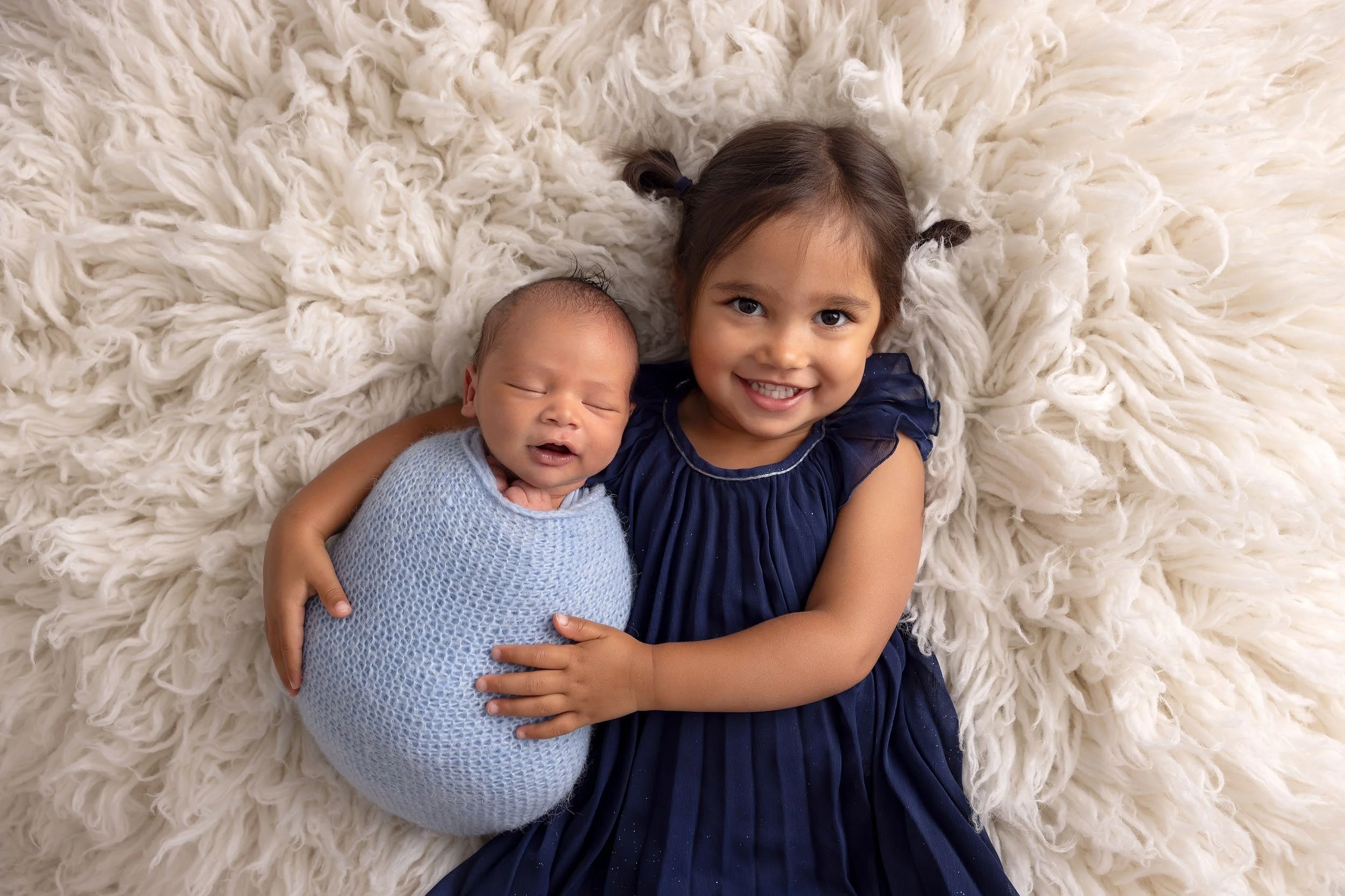A beautiful sibling photo of little girl with dark hair in pigtails smiling and holding a sleeping newborn wrapped in a blue blanket, lying on a fluffy cream-colored blanket.