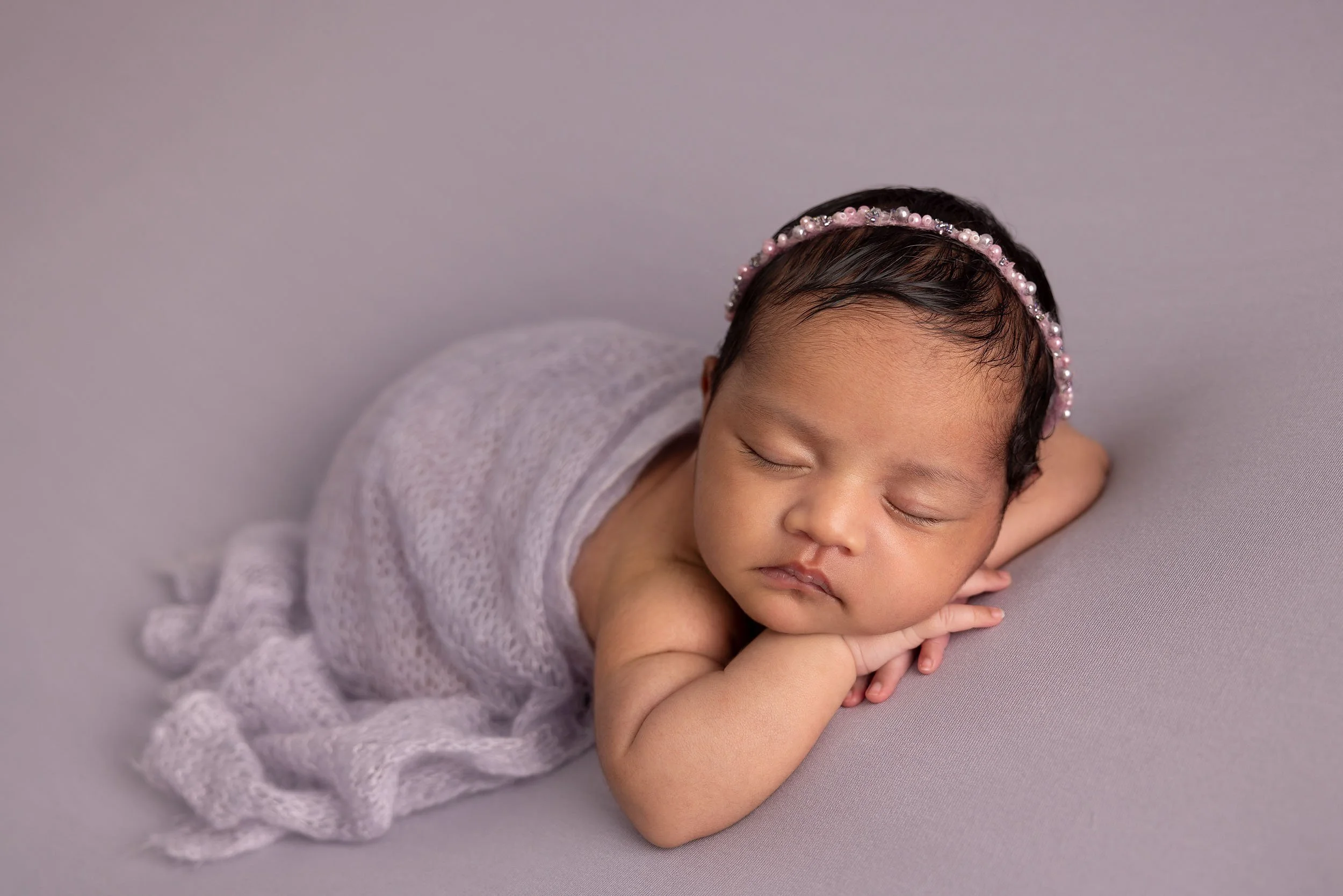A sleeping newborn baby with a pink pearl headband, resting on a soft gray surface with folded hands under her chin.