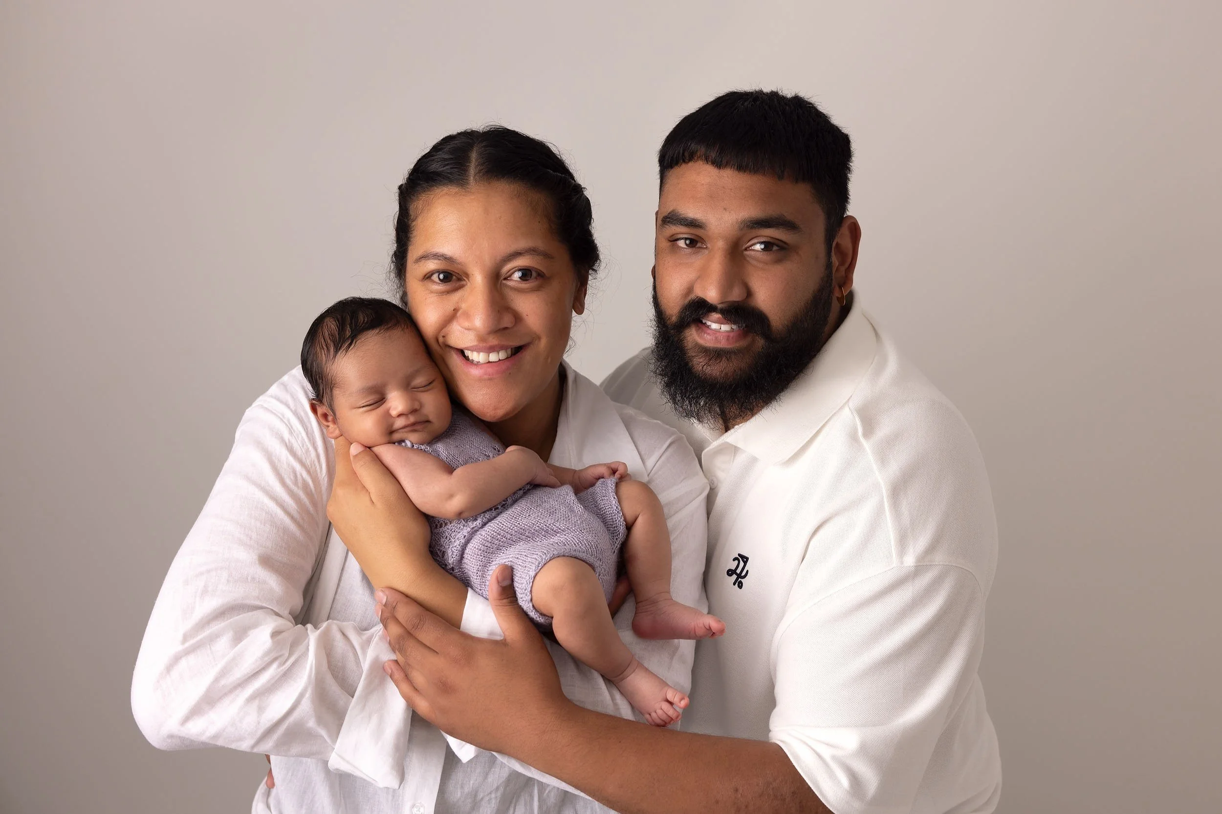 A happy family of three, an woman, man, and a newborn baby, posing together against a plain background. The woman is holding the baby, and all are smiling warmly at the camera.