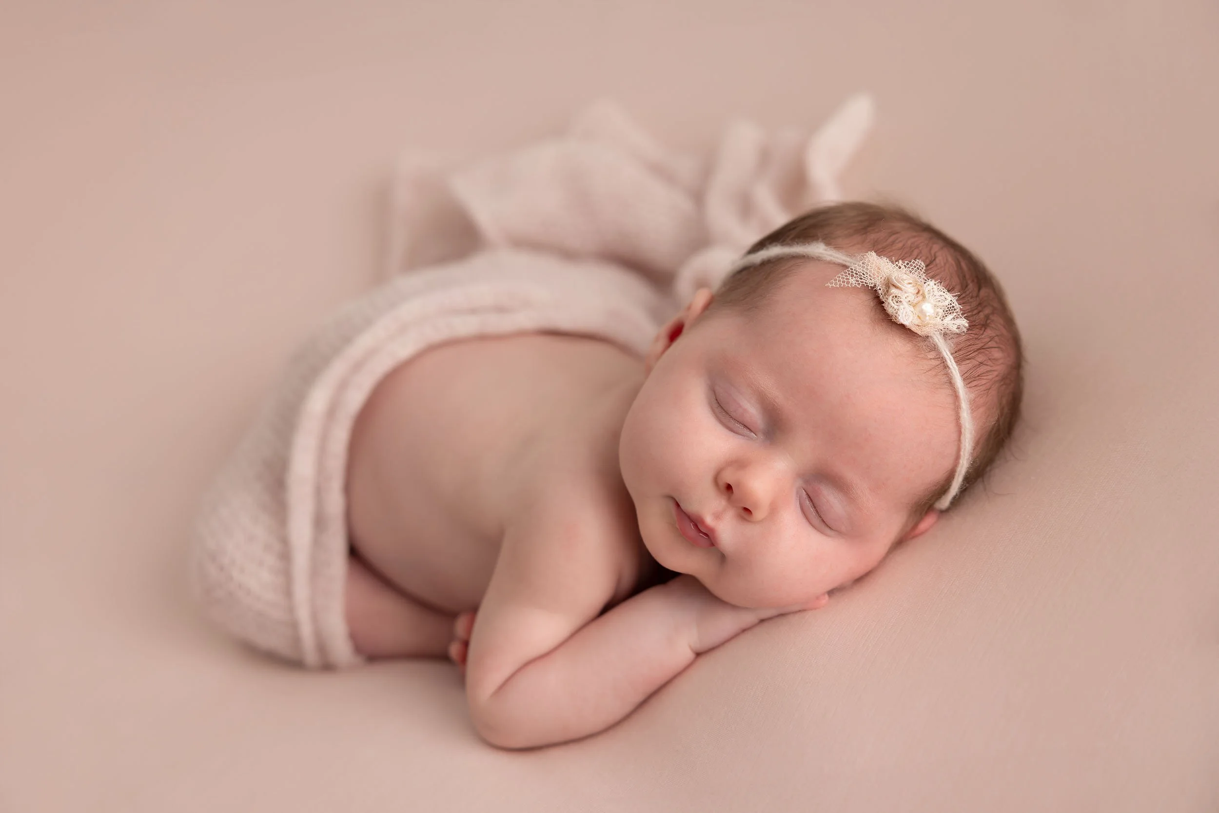 Sleeping baby girl with a headband, lying on her side on a soft beige surface, wrapped in a pink blanket.